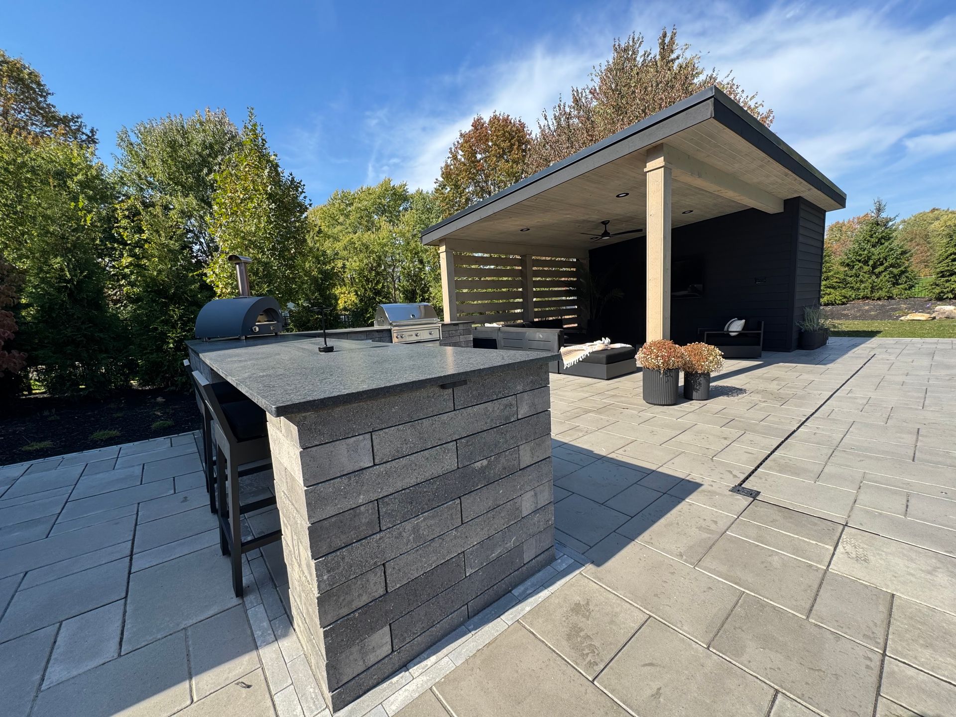 Outdoor kitchen with a brick countertop and pizza oven in a paved patio. A cozy, dark wood gazebo with seating and plants is set against a backdrop of lush trees.