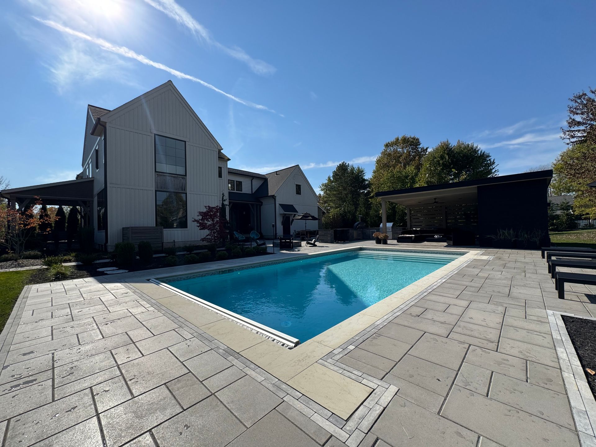 Modern house with a light exterior and large windows is next to a pristine blue pool. The spacious patio has lounge chairs and a shaded sitting area.