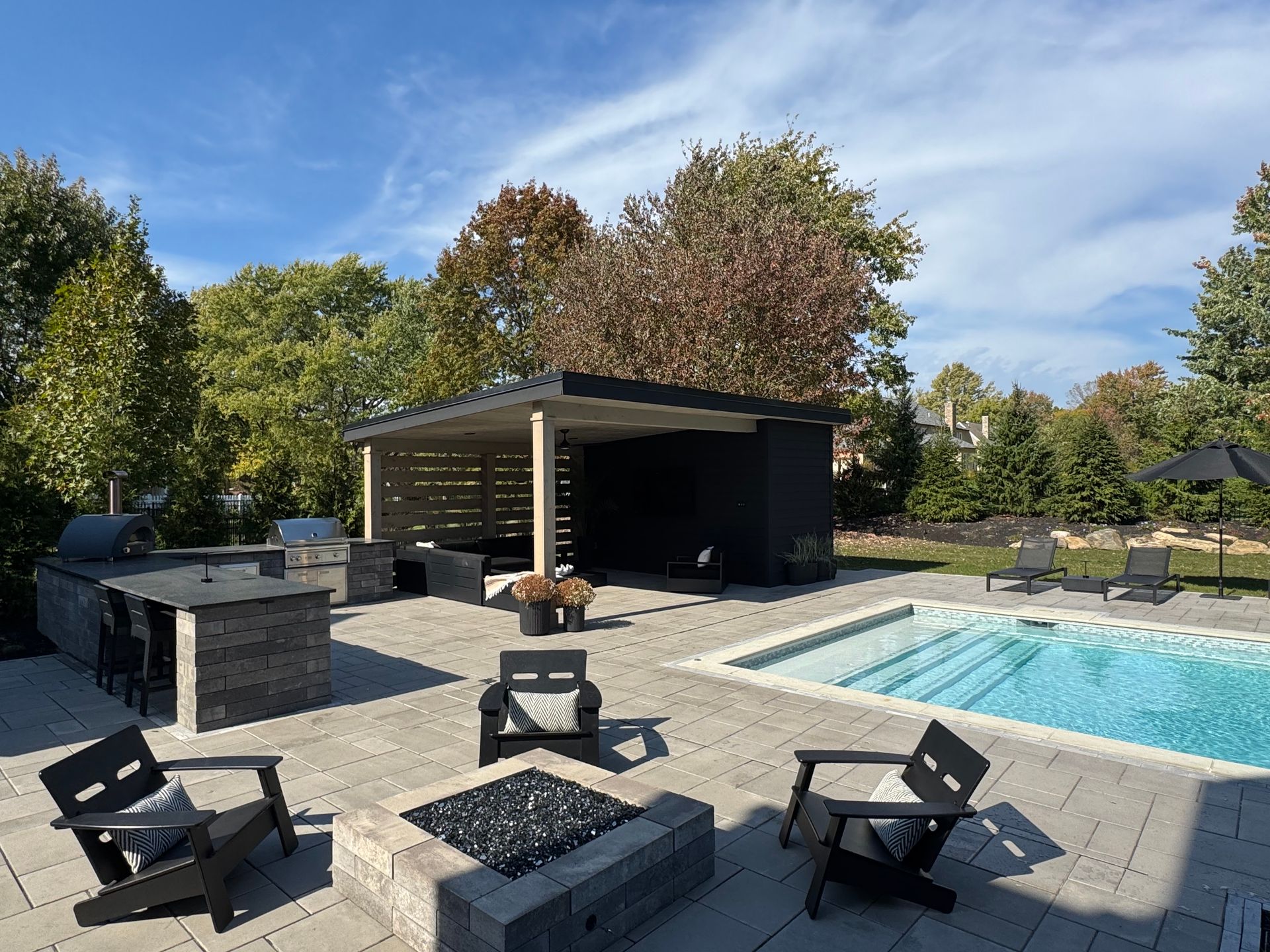Modern backyard with a sleek outdoor lounge by a rectangular pool. Features a fire pit, bar with grill, and pergola. Surrounded by lush trees under a bright sky.