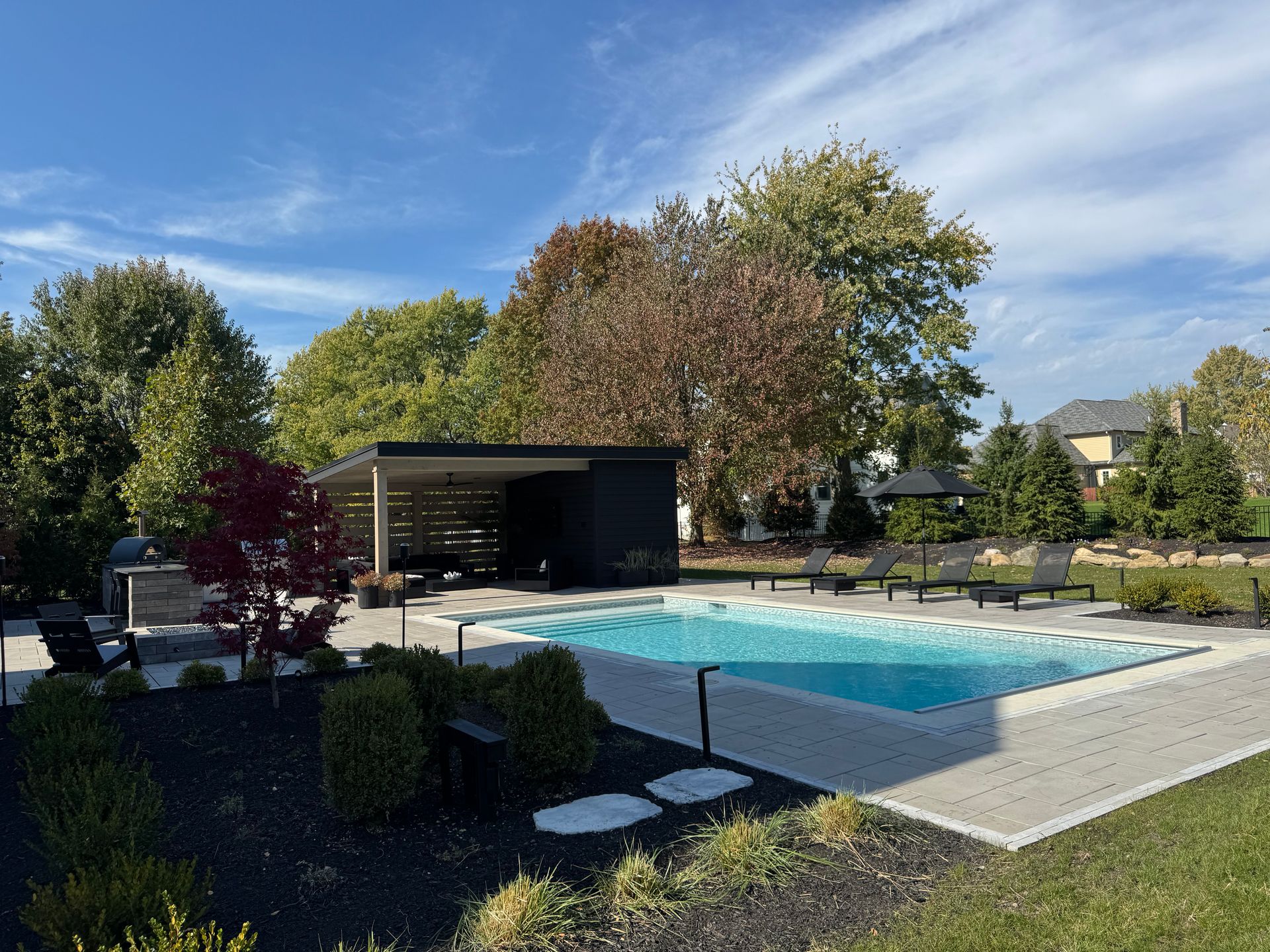 Modern backyard with a rectangular pool surrounded by patio tiles and lounge chairs. A pergola and barbecue area enhance the tranquil, sunny setting.