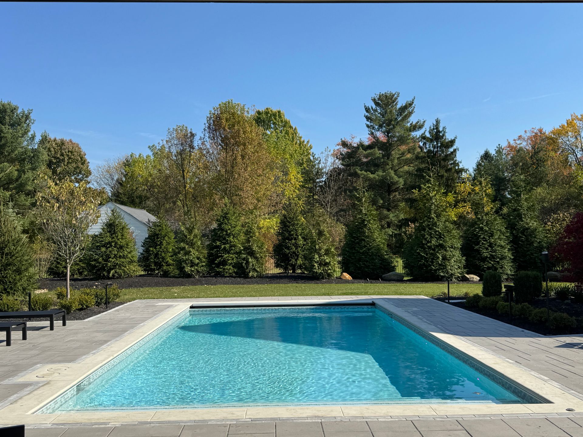 Rectangular swimming pool with clear, blue water, set in a spacious stone patio. Lush green trees and a clear blue sky surround the area, evoking a serene and inviting atmosphere.