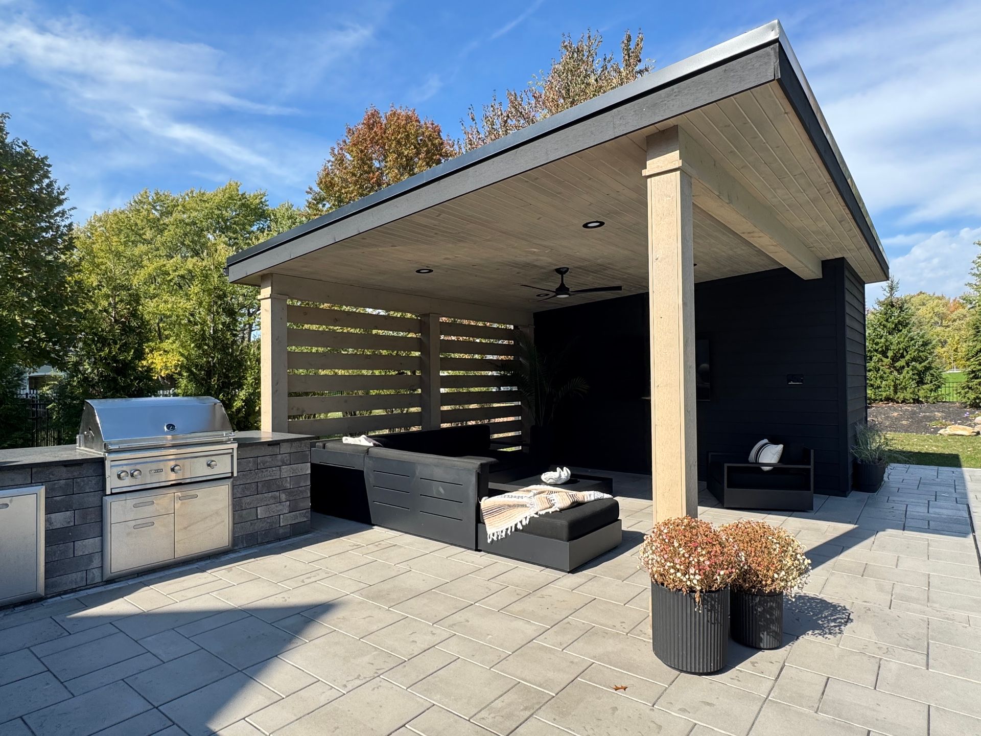 Modern outdoor patio with a wooden pergola, black furniture, and a stainless steel barbecue. Paved flooring and potted plants, under a bright blue sky.