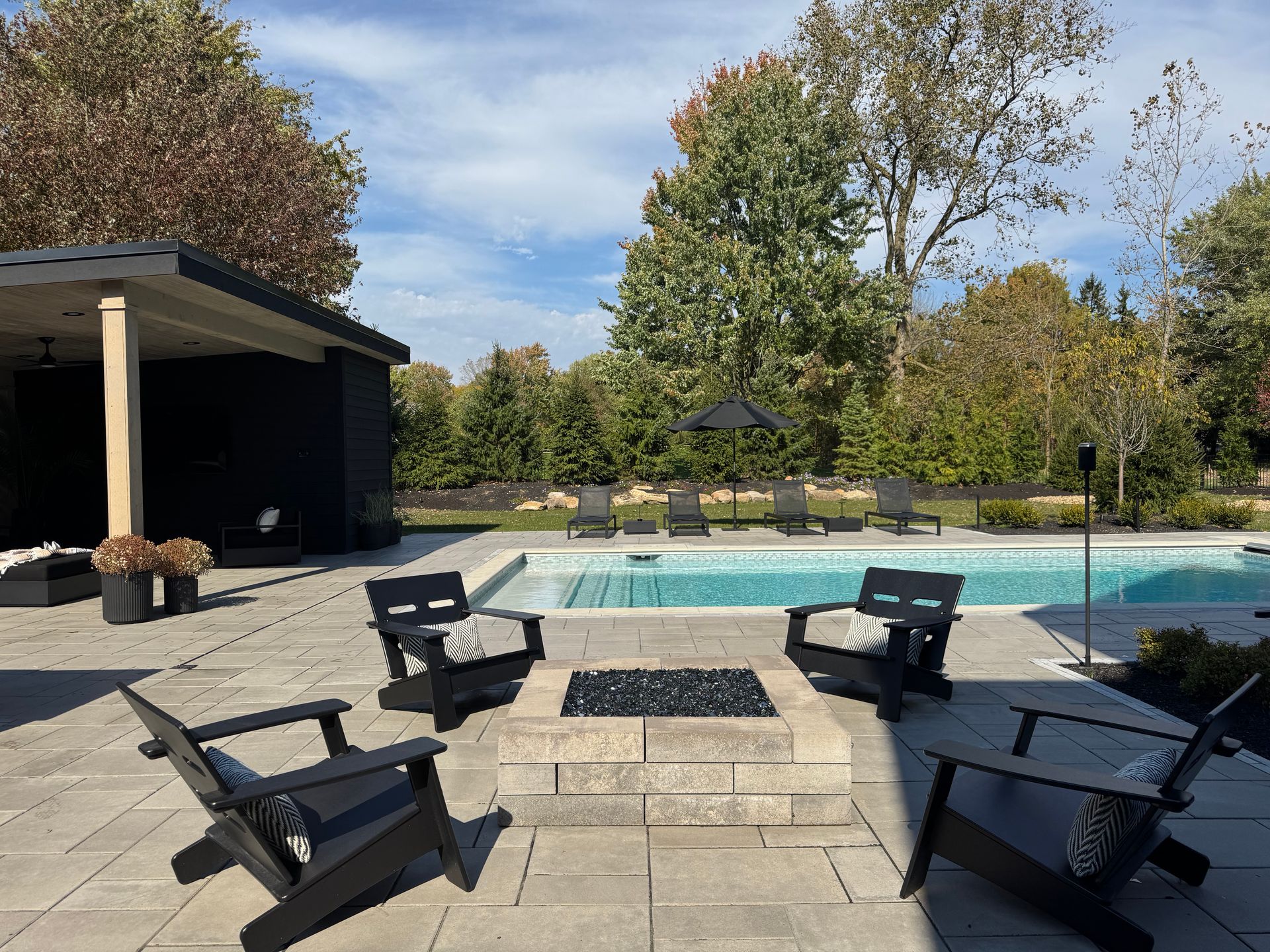 Modern backyard with a pool, surrounded by trees. Features black lounge chairs, a fire pit with black chairs, and a shaded sitting area. Relaxing atmosphere.