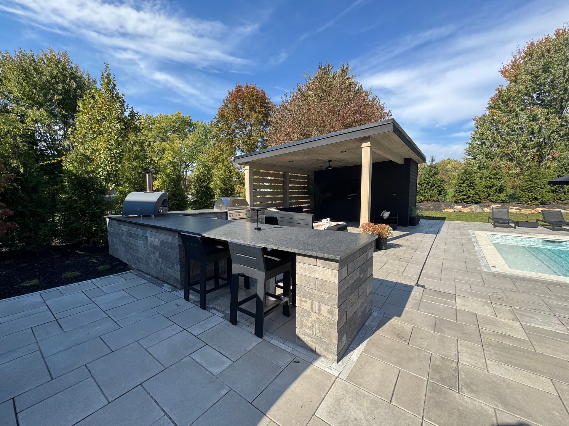 Outdoor kitchen with a stone counter and stools, adjacent to a covered patio with a grill. Surrounded by lush greenery and a pool under a sunny sky.