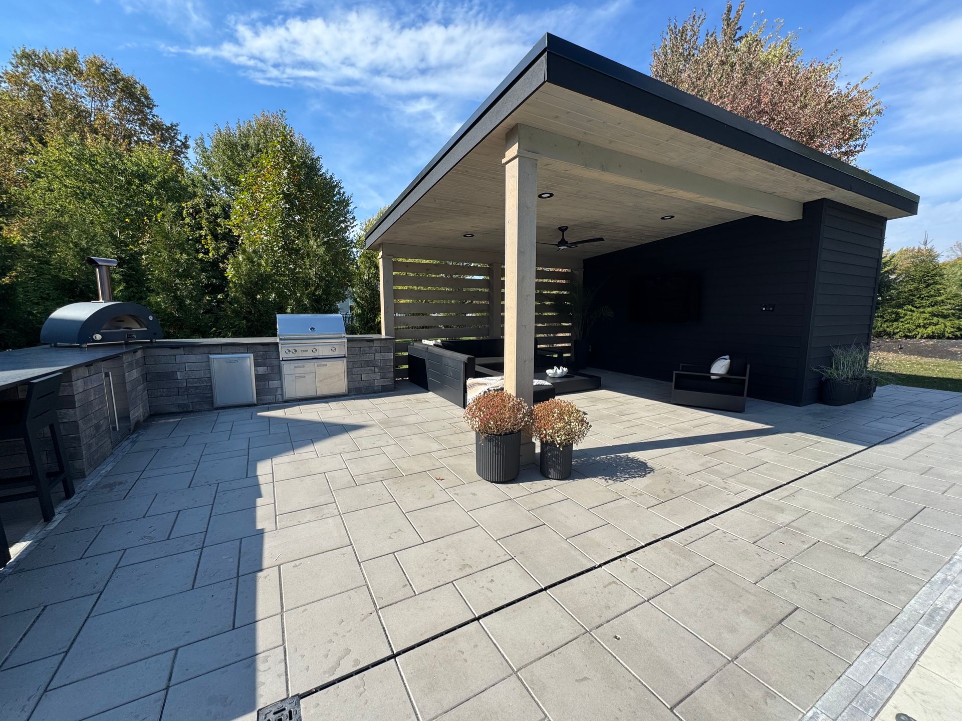 Modern outdoor patio with a sleek wood-fired pizza oven and grill. Covered seating area with a ceiling fan and cozy furniture. Bright, sunny day with a calm, inviting atmosphere.