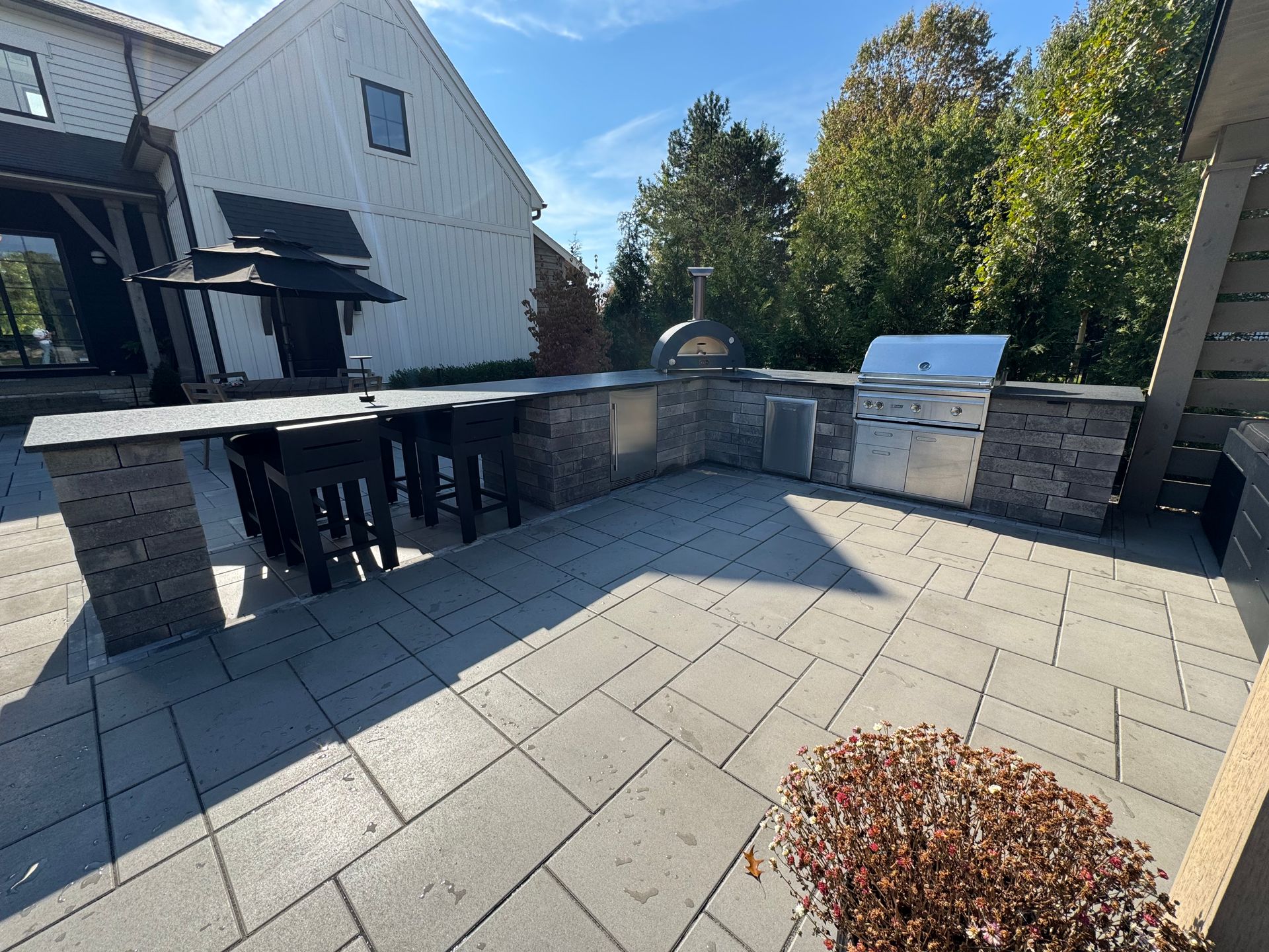 Outdoor kitchen with a stone counter, built-in grill, and pizza oven on a tiled patio. Nearby is a dining table with chairs. Trees provide a lush backdrop.