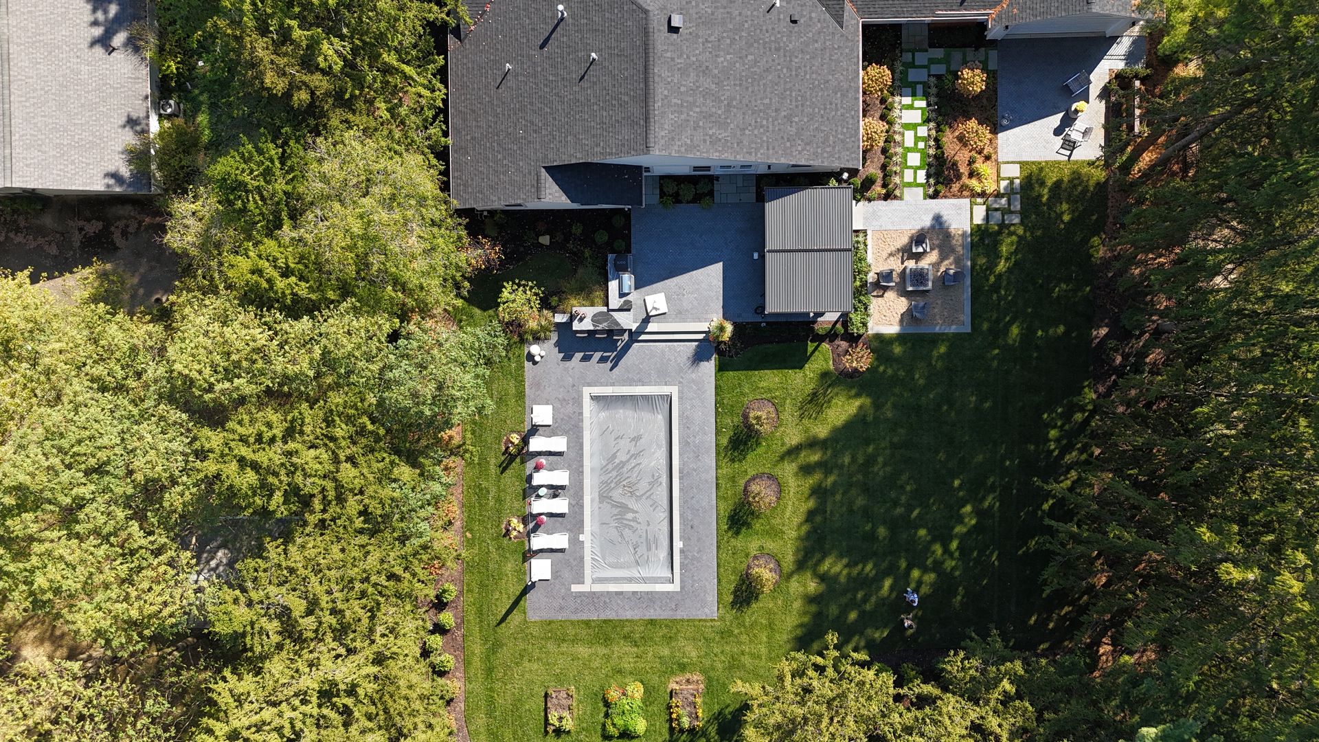 Aerial view of a house with a gray roof, a rectangular pool surrounded by lounge chairs, green lawn, trees, and a patio area with furniture.