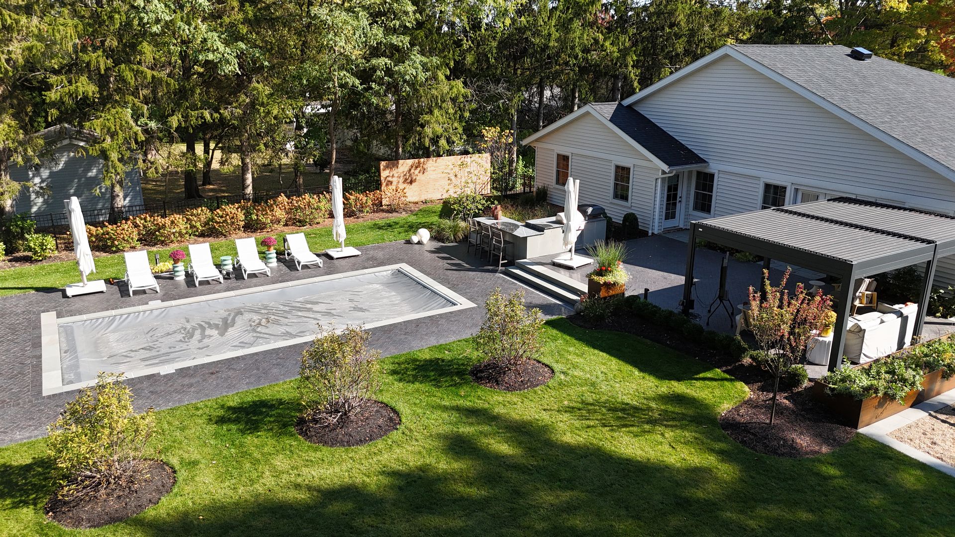 Aerial view of a modern backyard with a rectangular pool, sun loungers, white umbrellas, and a cozy patio next to a white house with a gray roof.