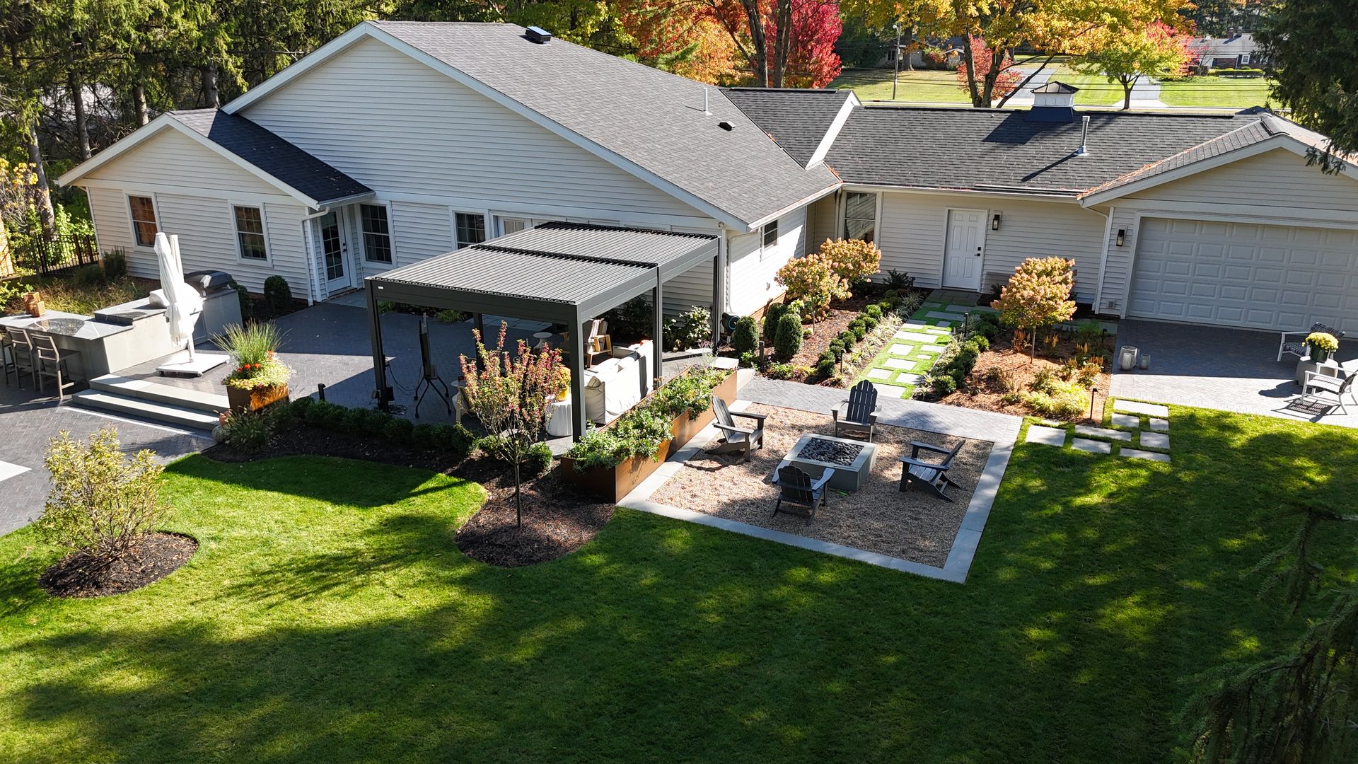 Aerial view of a house with a landscaped backyard. Features include a pergola, patio, fire pit, and well-maintained garden. Trees with autumn leaves provide a warm atmosphere.