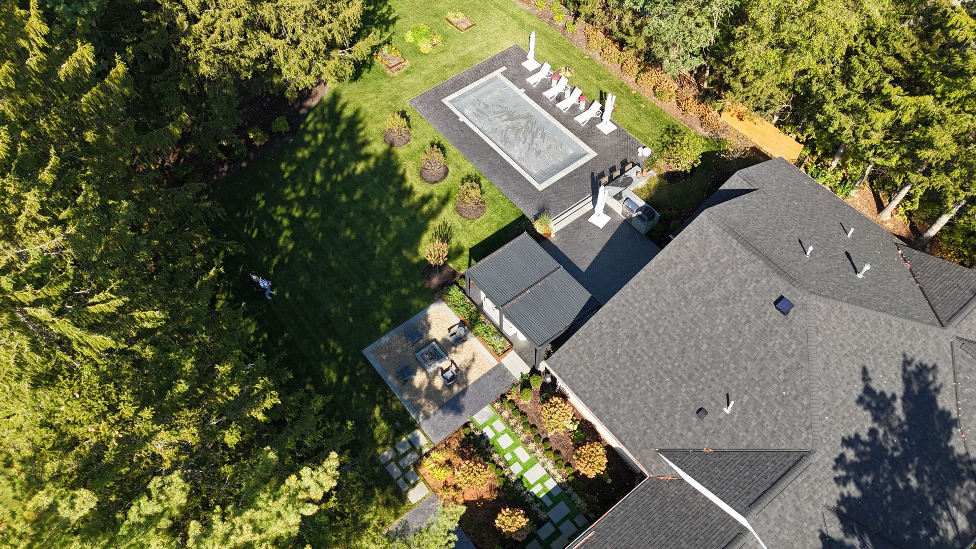 Aerial view of a backyard with a large swimming pool surrounded by lounge chairs, greenery, and tall trees. Adjacent is a house with a gray roof.