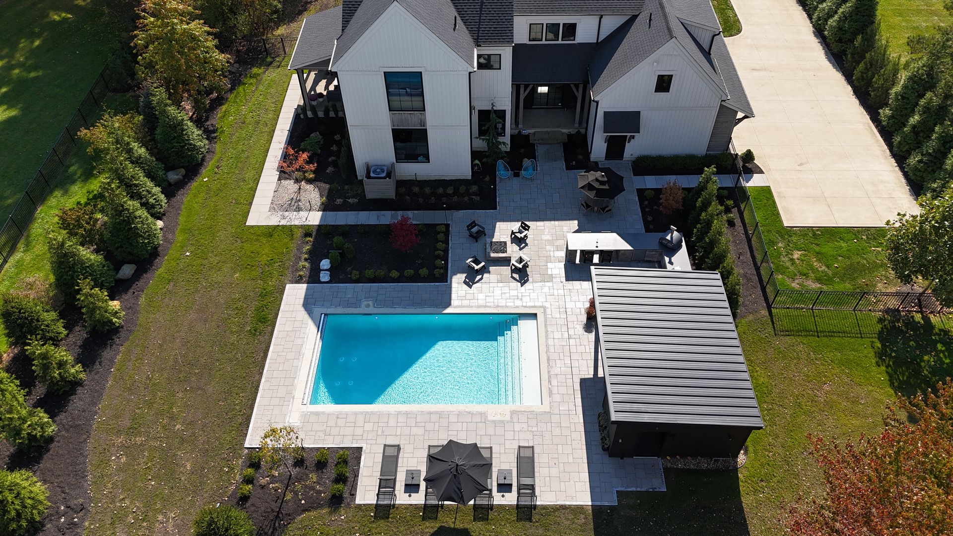 Aerial view of a modern backyard featuring a rectangular swimming pool with light blue water, surrounded by beige stone tile. There's a patio with chairs and a small wooden structure nearby.