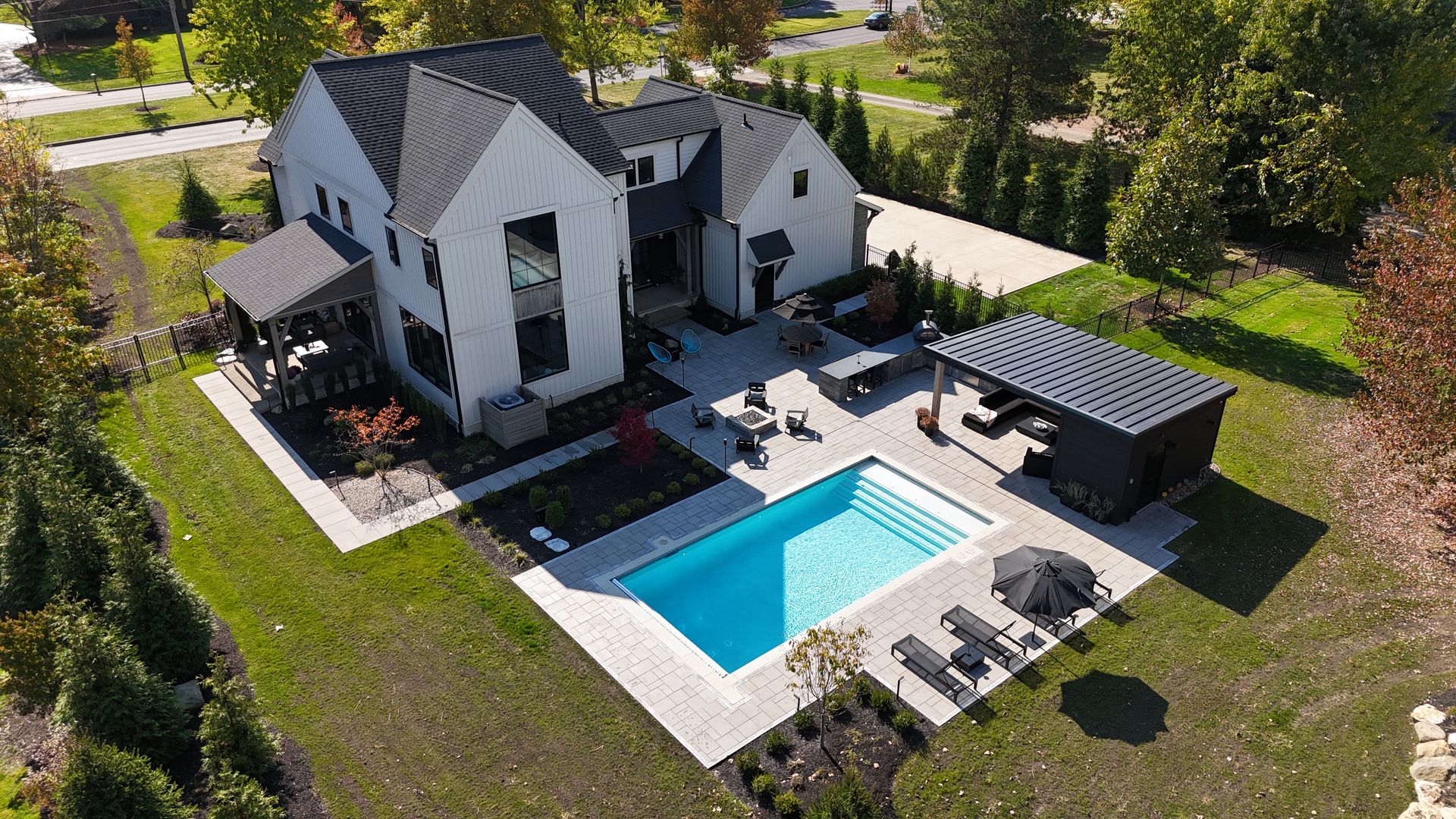 Aerial view of a modern white house with a black roof. The backyard features a rectangular pool, patio furniture, and a stylish poolside cabana, surrounded by green trees.