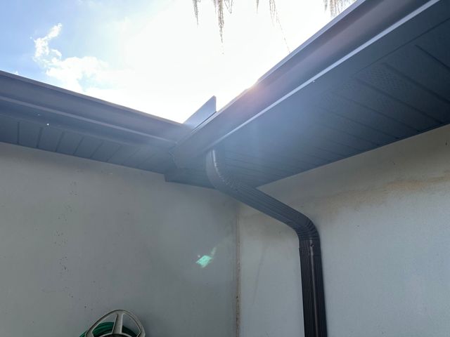 Sunlit corner of a house roof with dark gutters and downspout against a white wall Sunlit corner of a house roof with dark gutters and downspout against a white wall