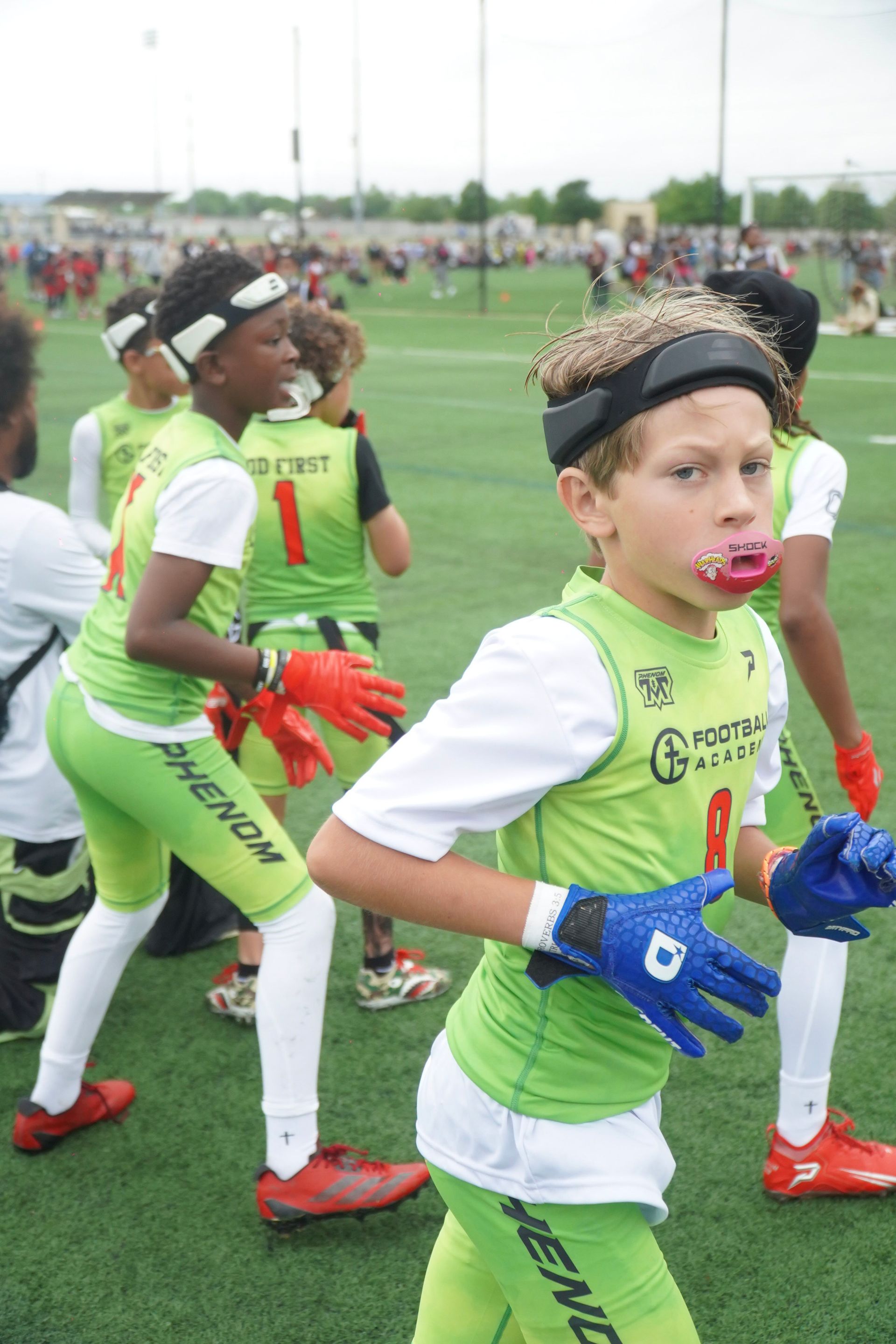 Young athletes in green and white uniforms run on a grassy field during a sports event.