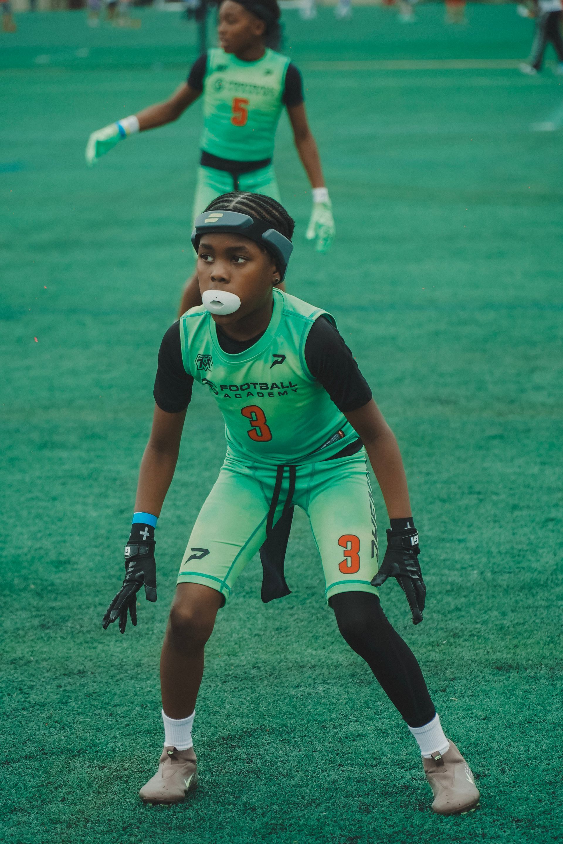 Youth lacrosse player in teal uniform crouches ready on a green field, with another player blurred behind.
