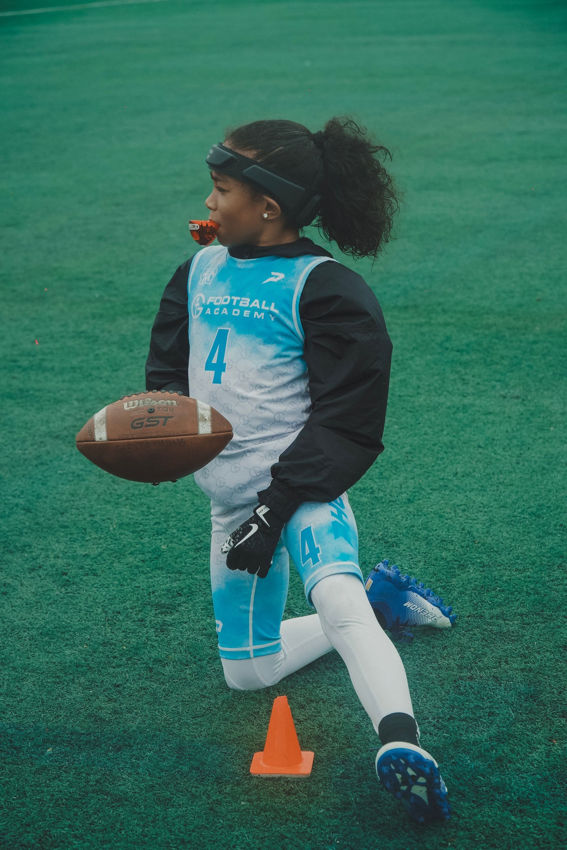 Child in blue football uniform holding a football beside an orange cone on a green turf field.