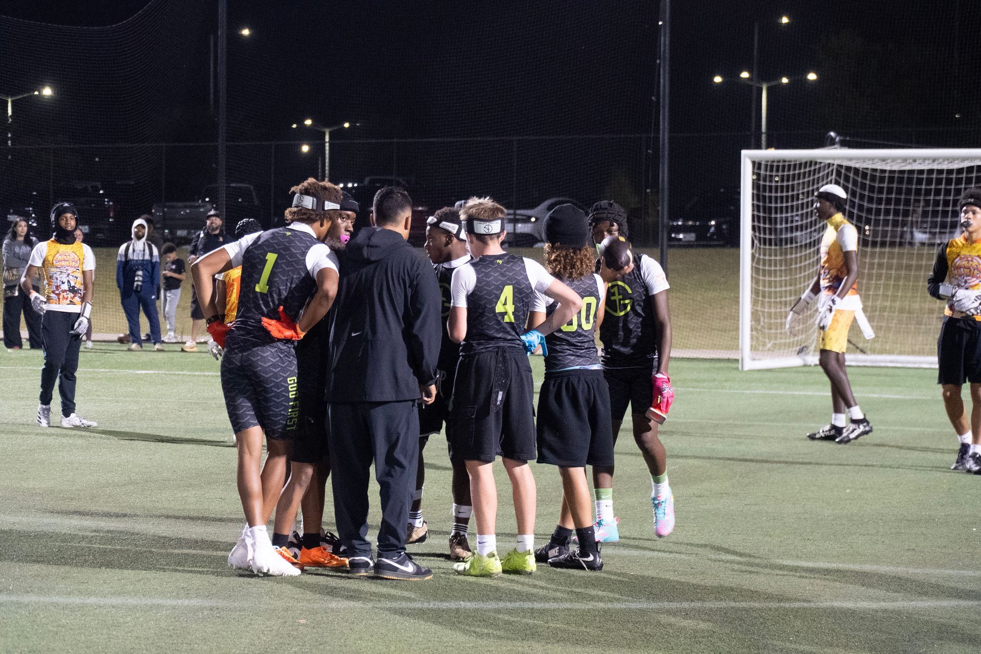 Soccer players huddle on an outdoor field at night near the goal, with a referee standing beside them.