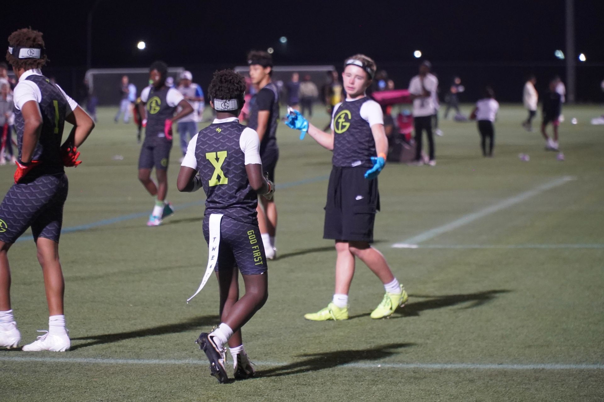 Flag football players on a lit field at night, one in black jersey with neon green “8” holding a flag.