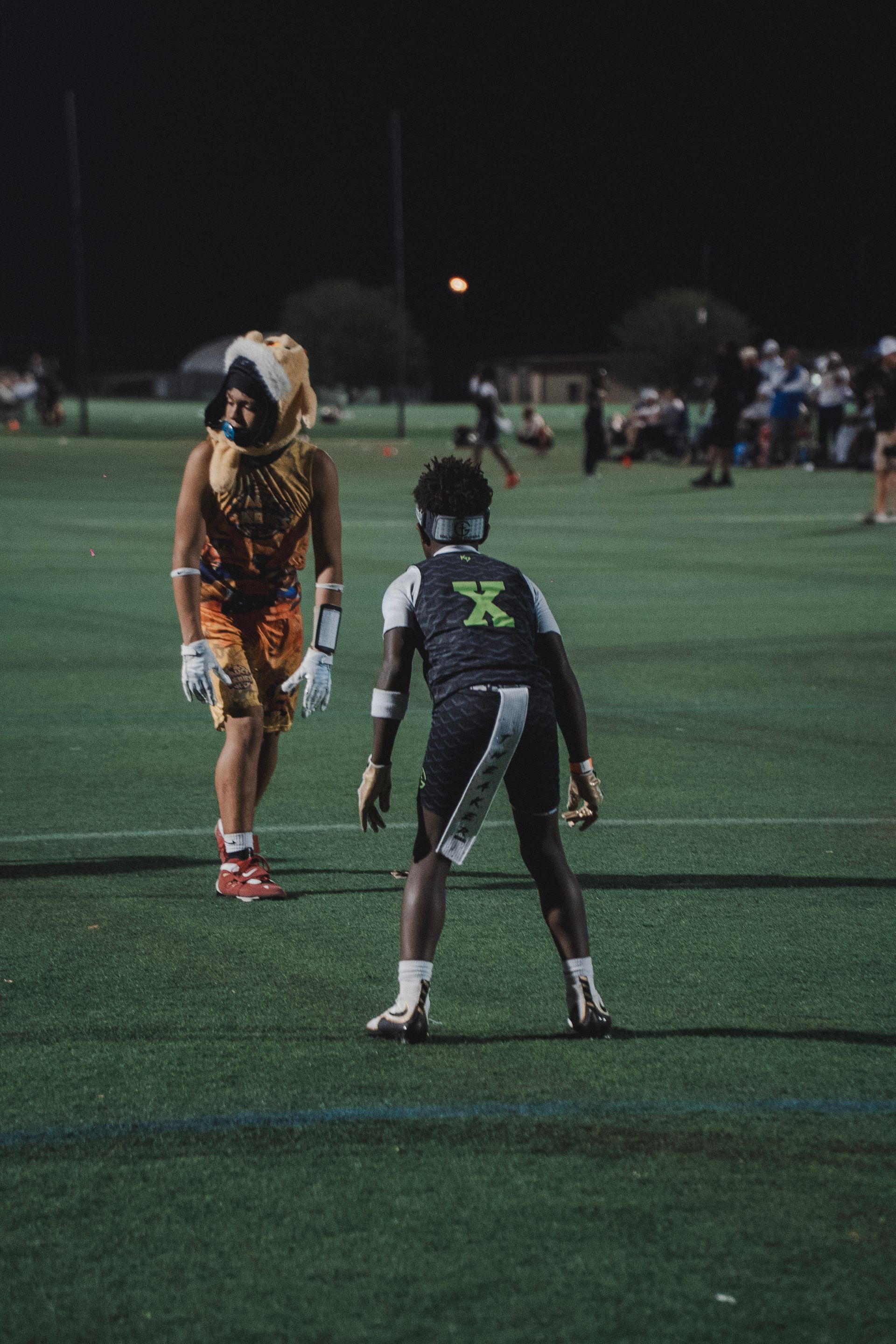 Two youth athletes face off on a lit field at night, one in black, one in orange and white.