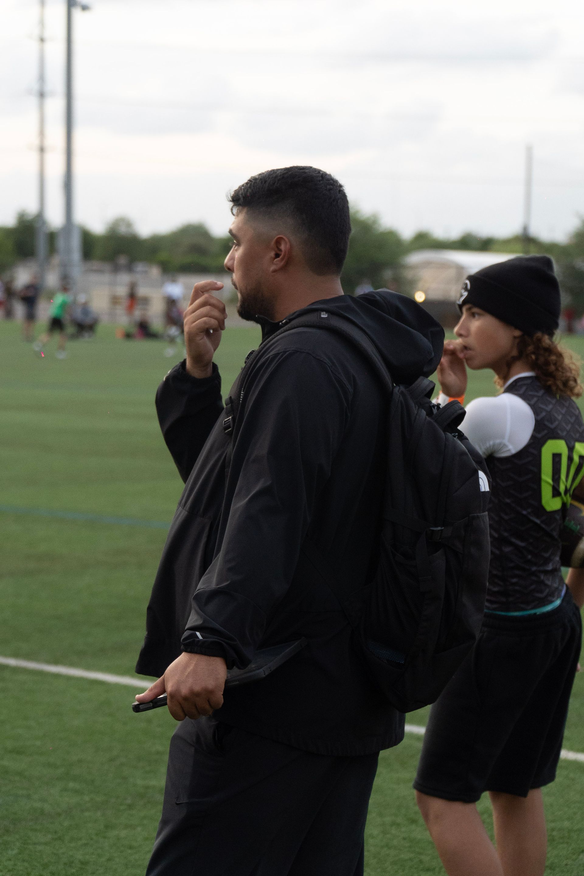 Two people in black sportswear standing on a grassy field, one holding a whistle and a phone.