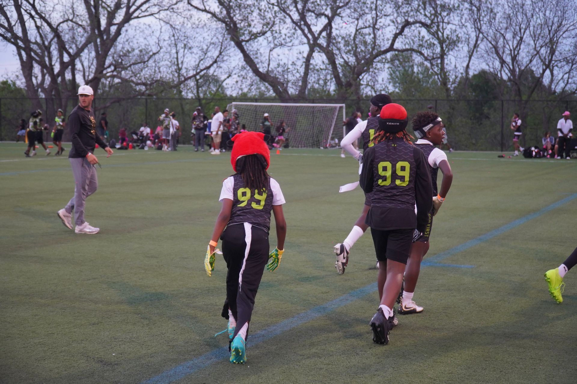 Youth flag football players in black uniforms and red helmets running on a grassy field near a goal