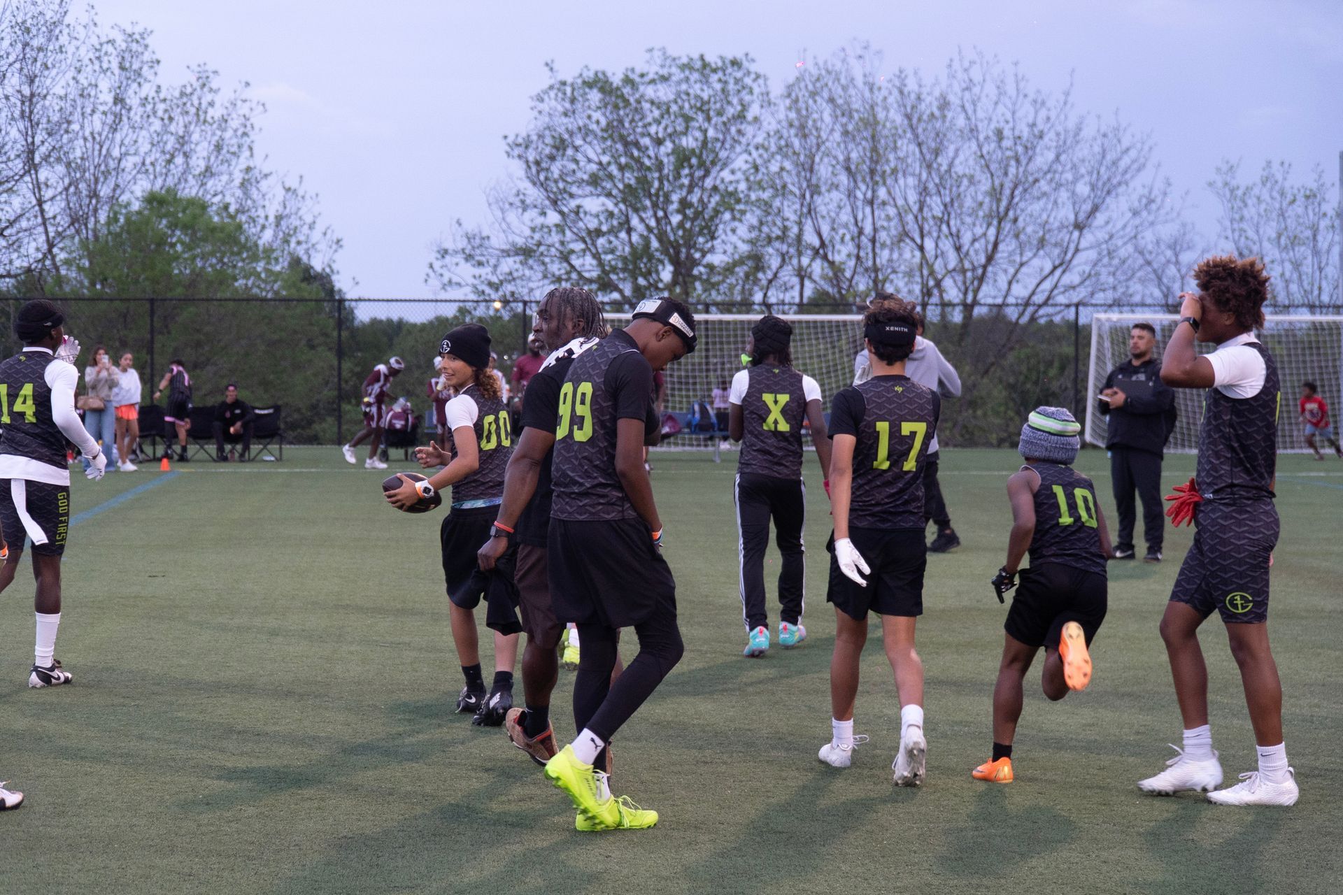 Football players in black jerseys celebrate on a grassy field, with trees and spectators in the background.