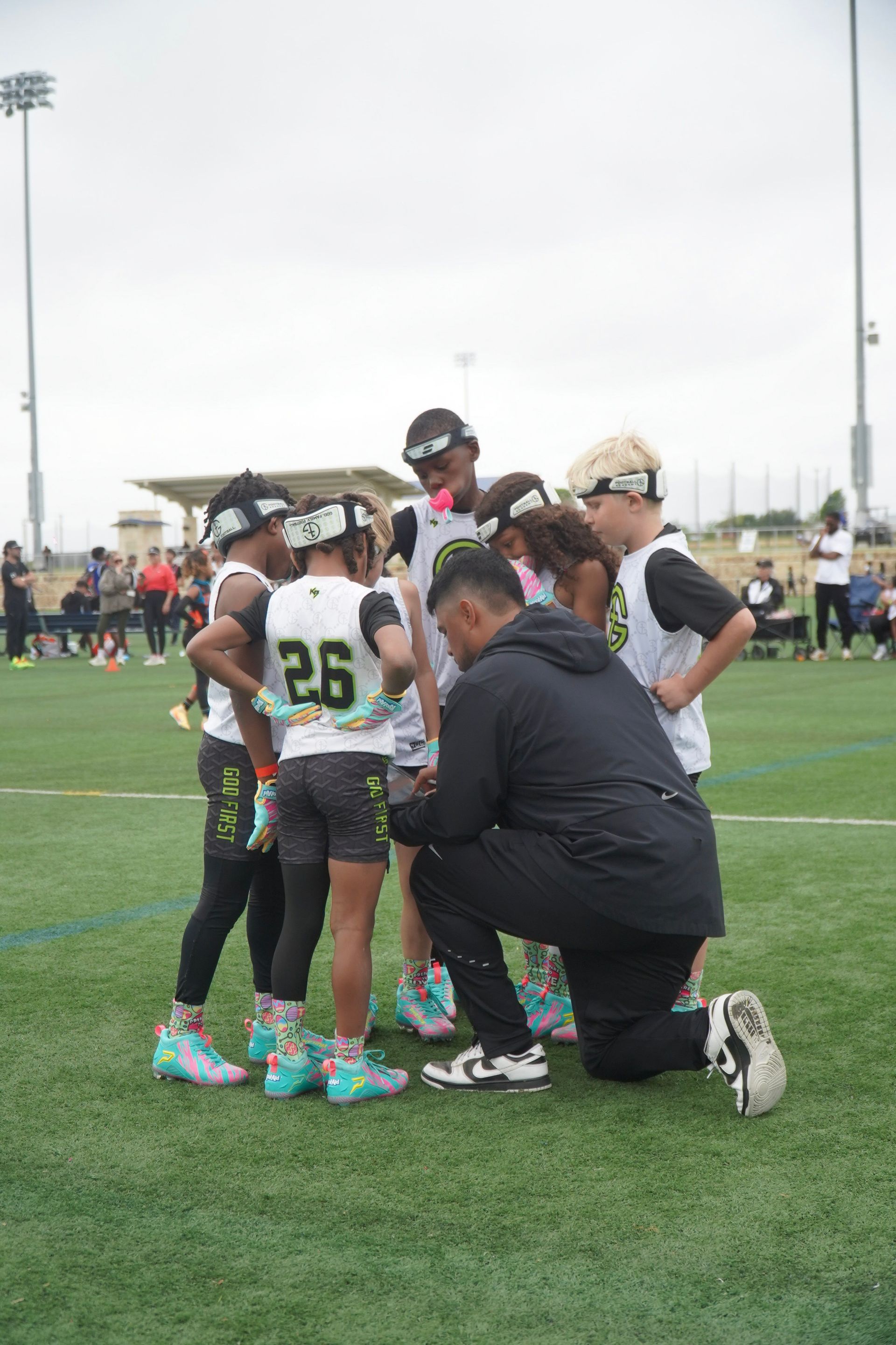 Youth lacrosse players huddle on a field while a coach kneels beside them.
