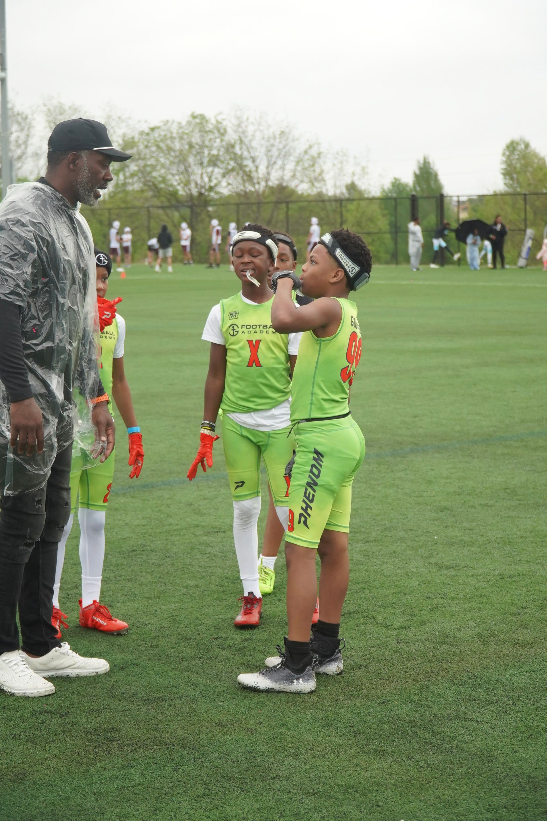 Youth lacrosse players in neon green uniforms huddle on a grassy field with a coach.