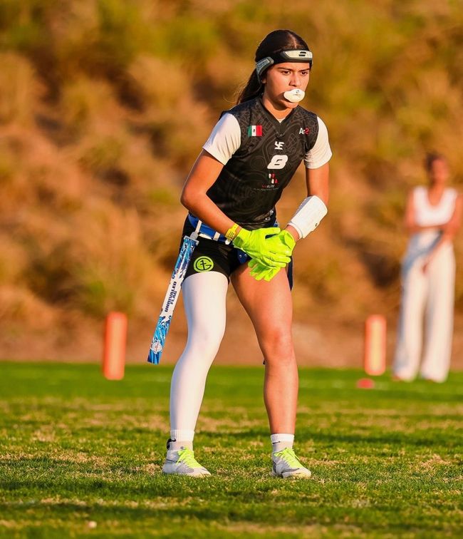 Athlete in black sportswear and neon shorts stands on a grassy field holding a hockey stick.