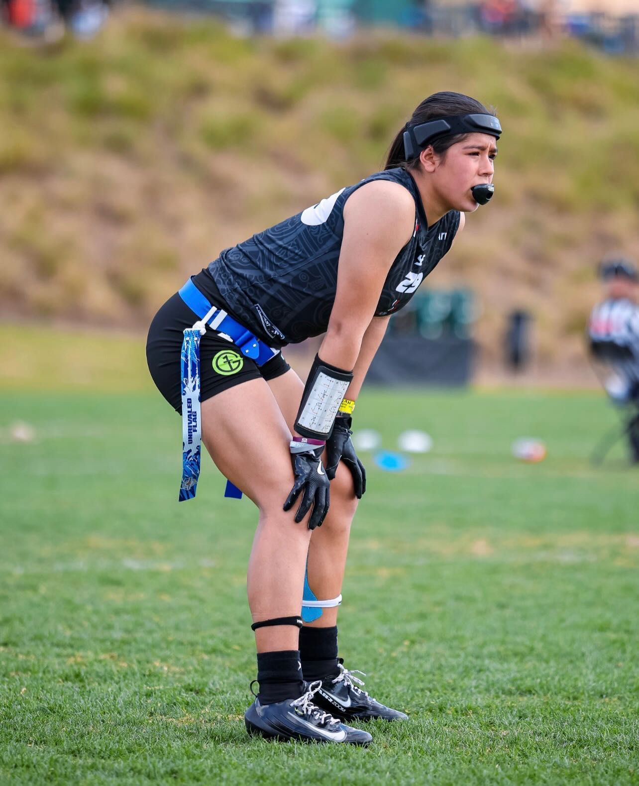 Flag football player in navy gear bent over on a grassy field, hands on knees during a game