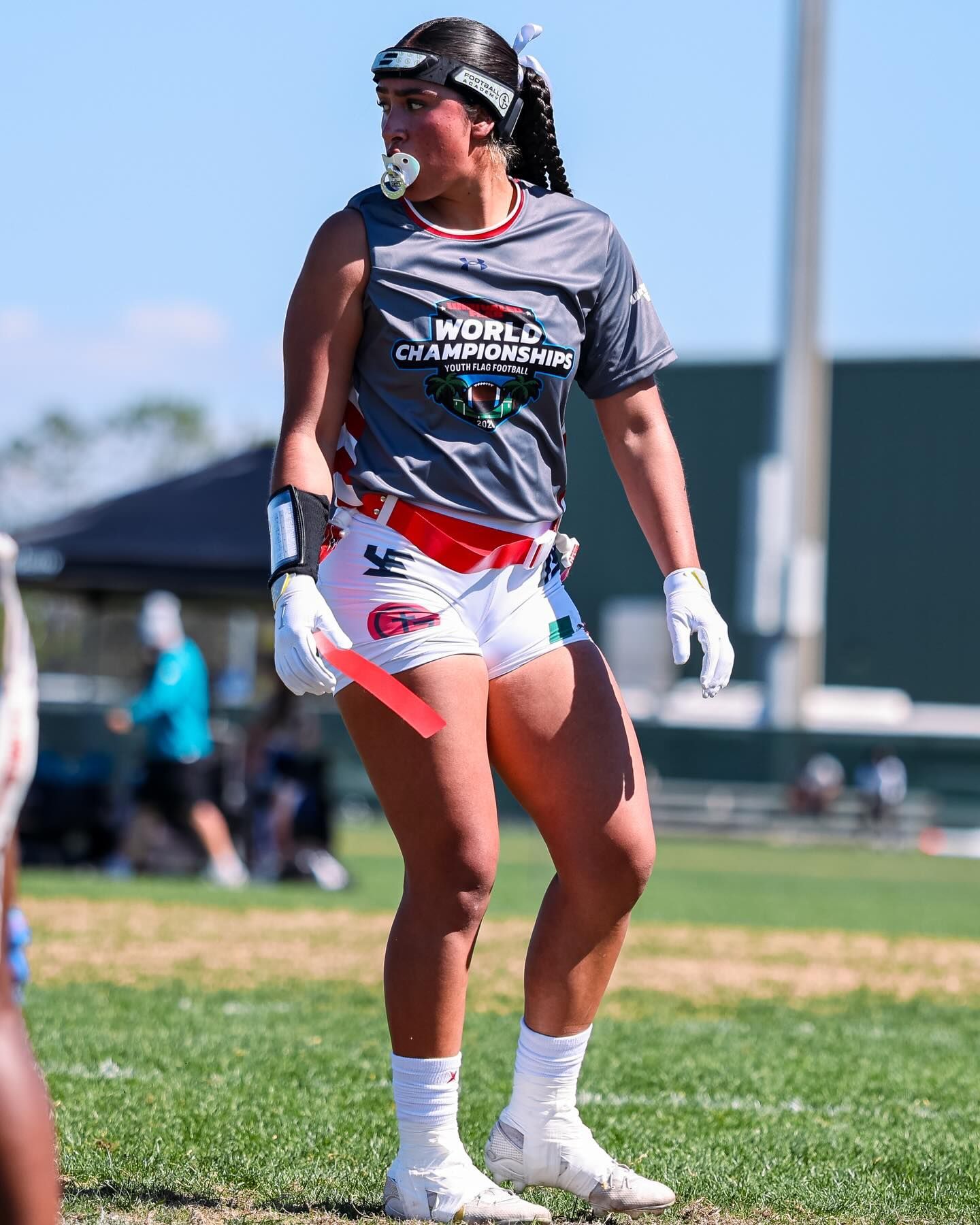 Female lacrosse player in gray and white uniform standing on grass field with helmet and gloves