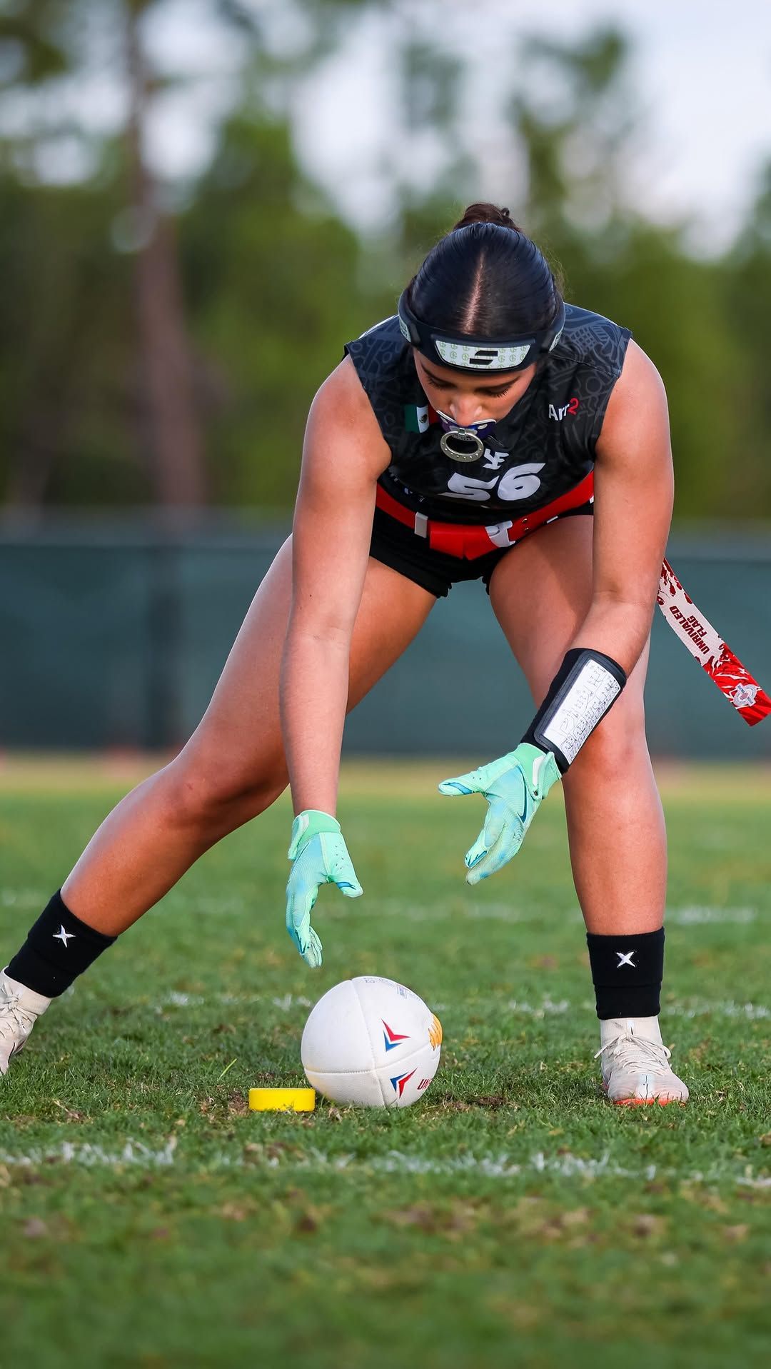 Field hockey player in black uniform reaching for a white ball on grass, wearing teal gloves and helmet.