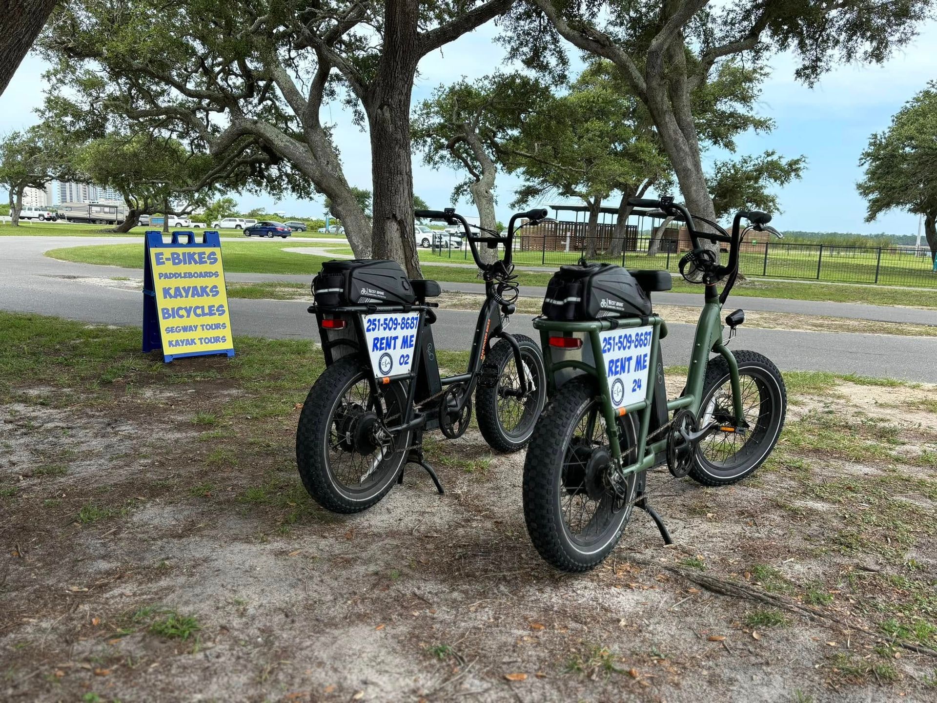 Two green electric bikes parked near a sign under trees on a grassy area.