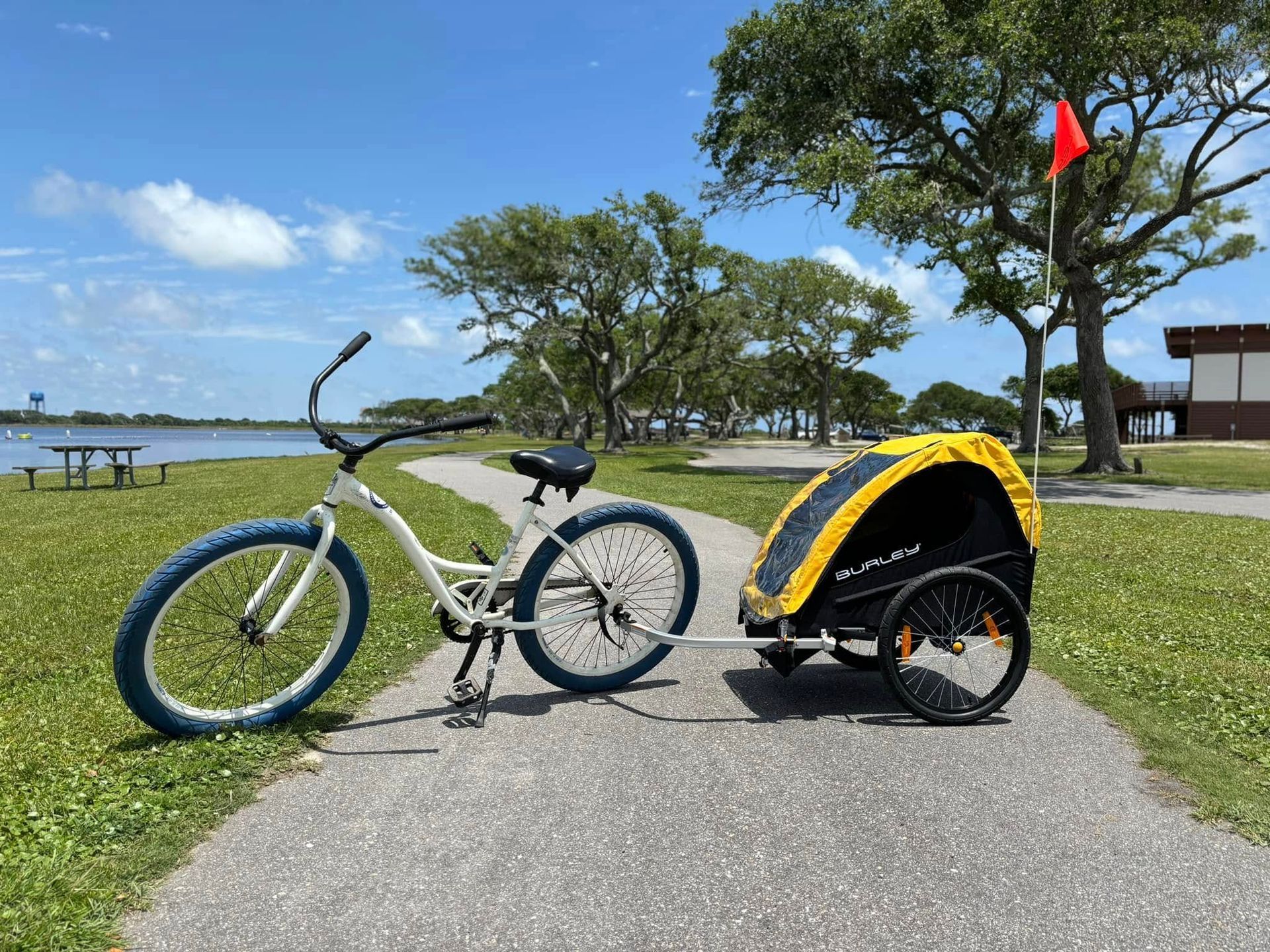 White bicycle with blue tires pulling a yellow trailer on a paved path near a body of water under a blue sky.