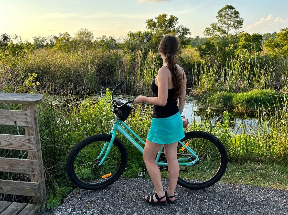 Woman standing by a turquoise bicycle, looking at a swampy area with green foliage, at sunset.