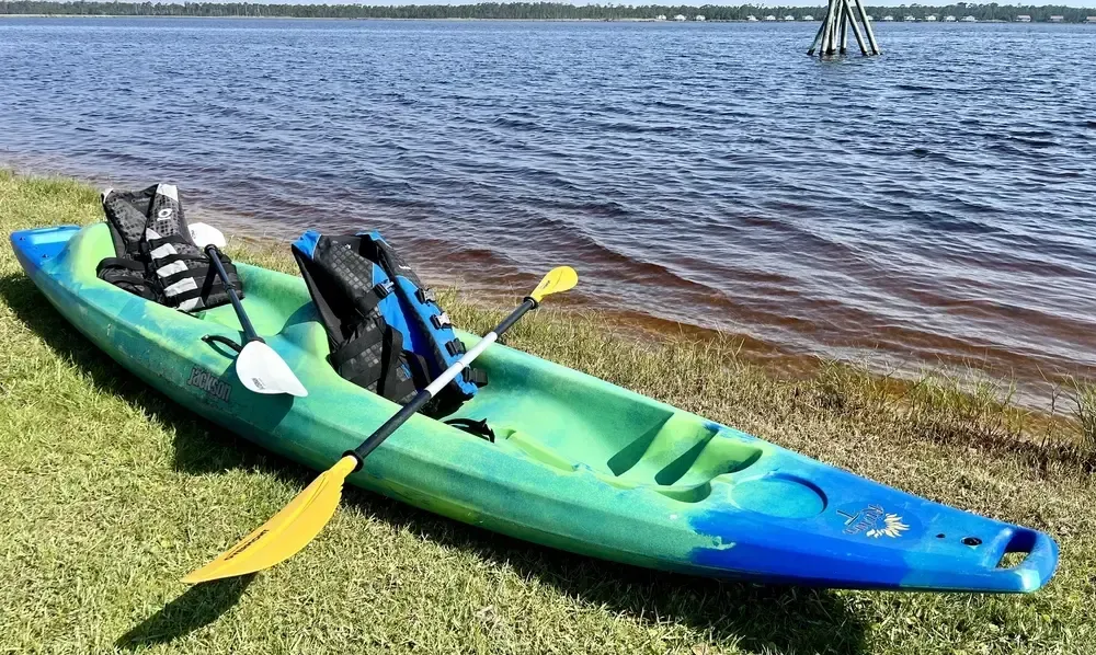 Green and blue kayak on a grassy shore next to a body of water, with life vests and paddle.