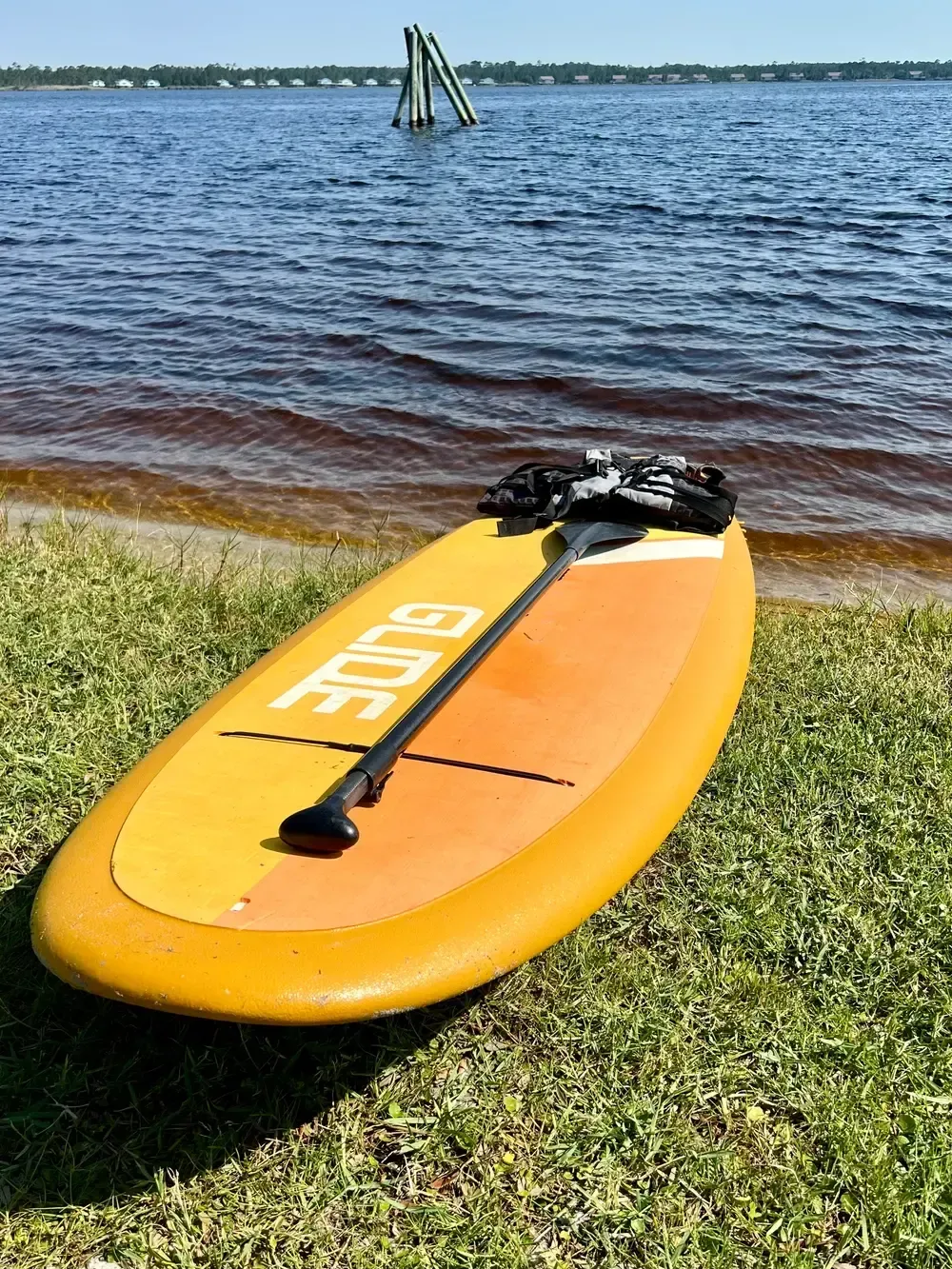 Yellow and orange paddleboard on grassy shore with paddle; water and sky in background.