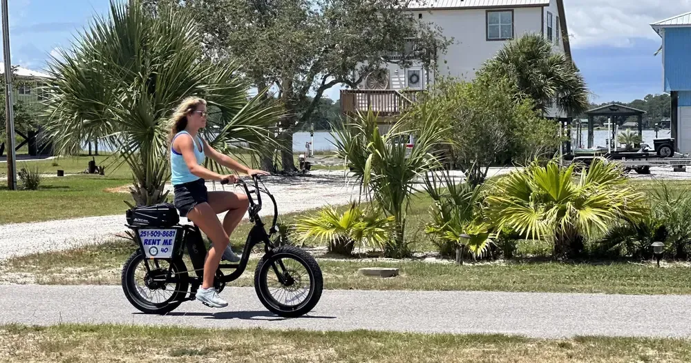 Woman rides a black electric bicycle on a paved path near buildings and foliage.