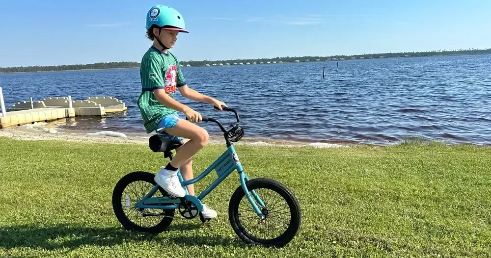 Boy riding a teal bicycle by a lake, wearing a helmet and summer clothes.