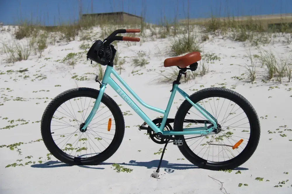 Light blue beach cruiser bicycle on white sand, with a brown seat and handlebars, under a clear sky.