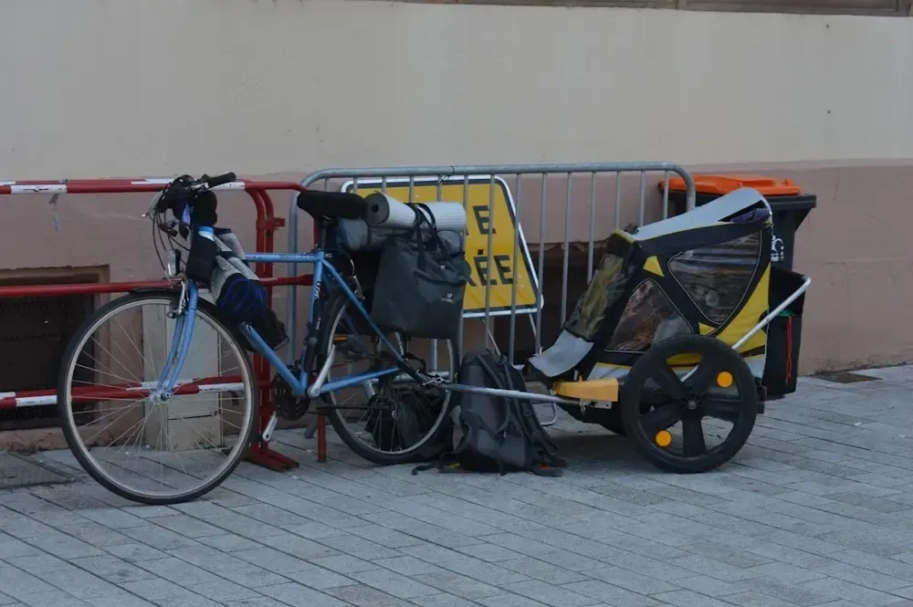 Blue bicycle with trailer carrying gear, parked in front of metal barriers and a building.