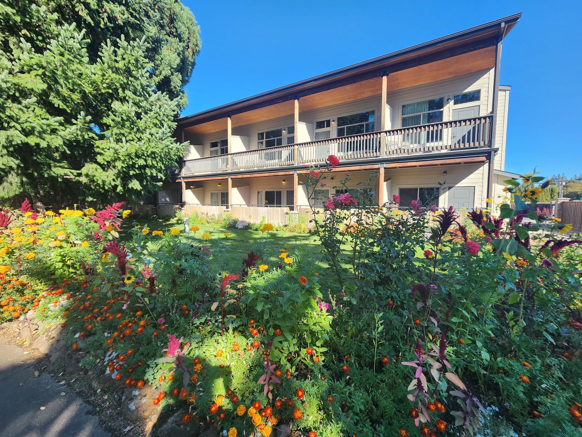Two-story building with balconies, surrounded by flowers and greenery, under a blue sky.