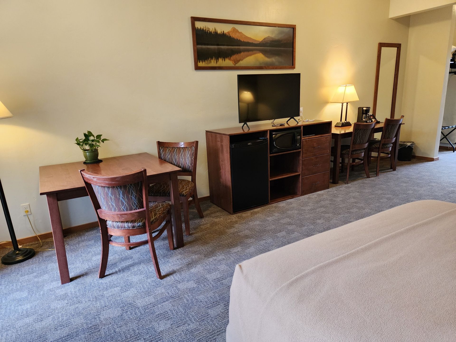 A hotel room with a bed, desk, chairs, TV, and dining area. Neutral colors with light carpet.
