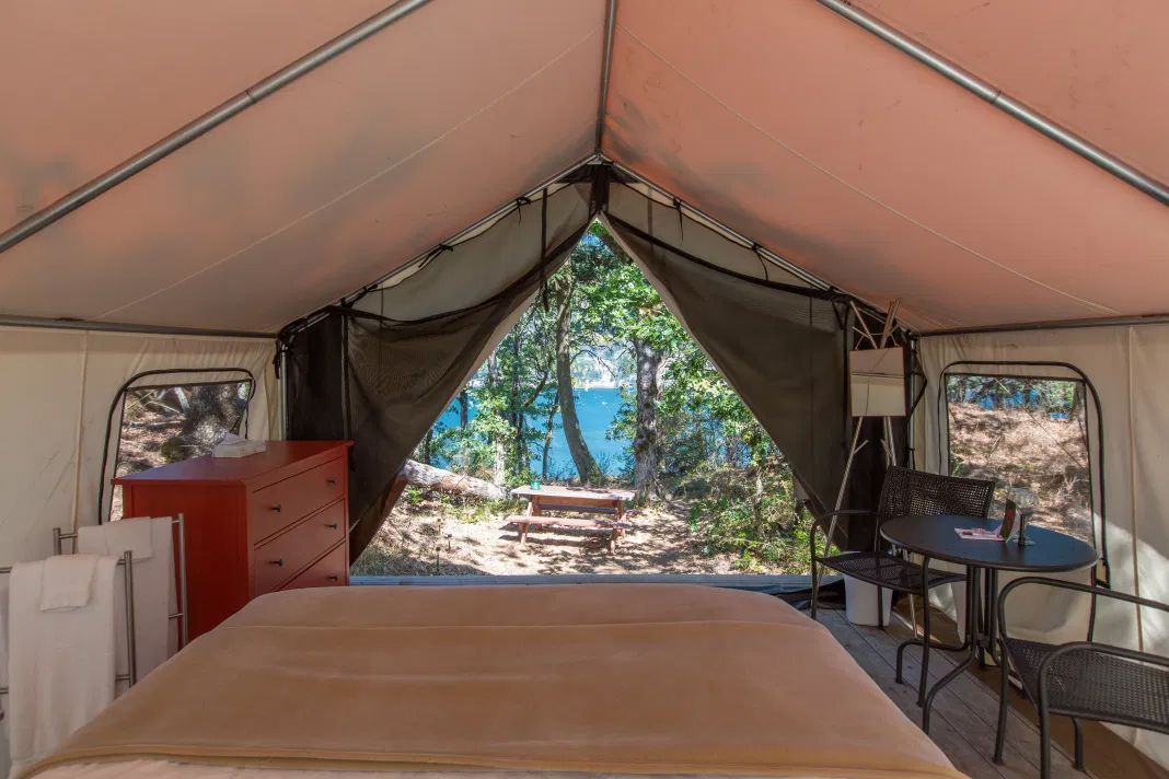 Interior of a glamping tent with bed, dresser, table, and view of water. Beige tent fabric.