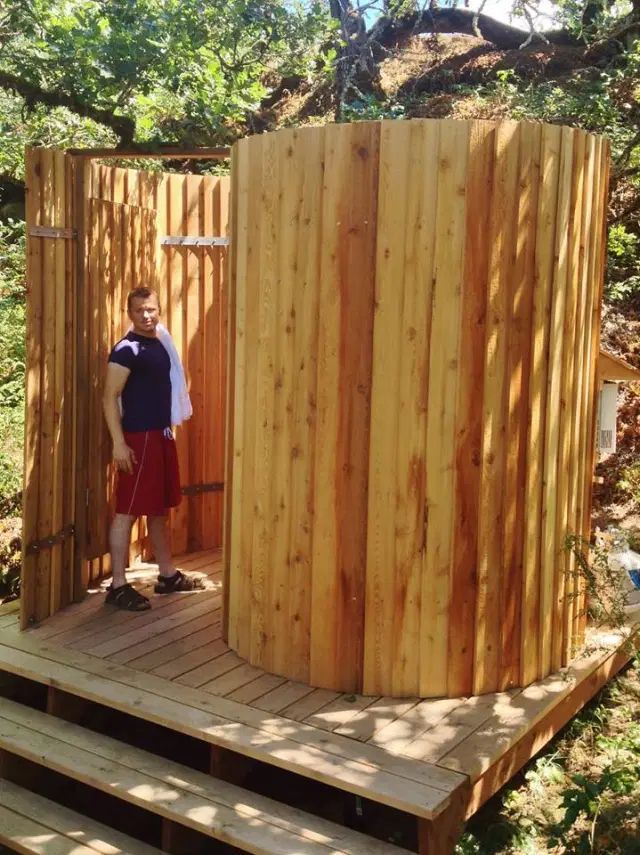 Man standing by a wooden outdoor shower on a deck, holding a towel. Green trees in background.