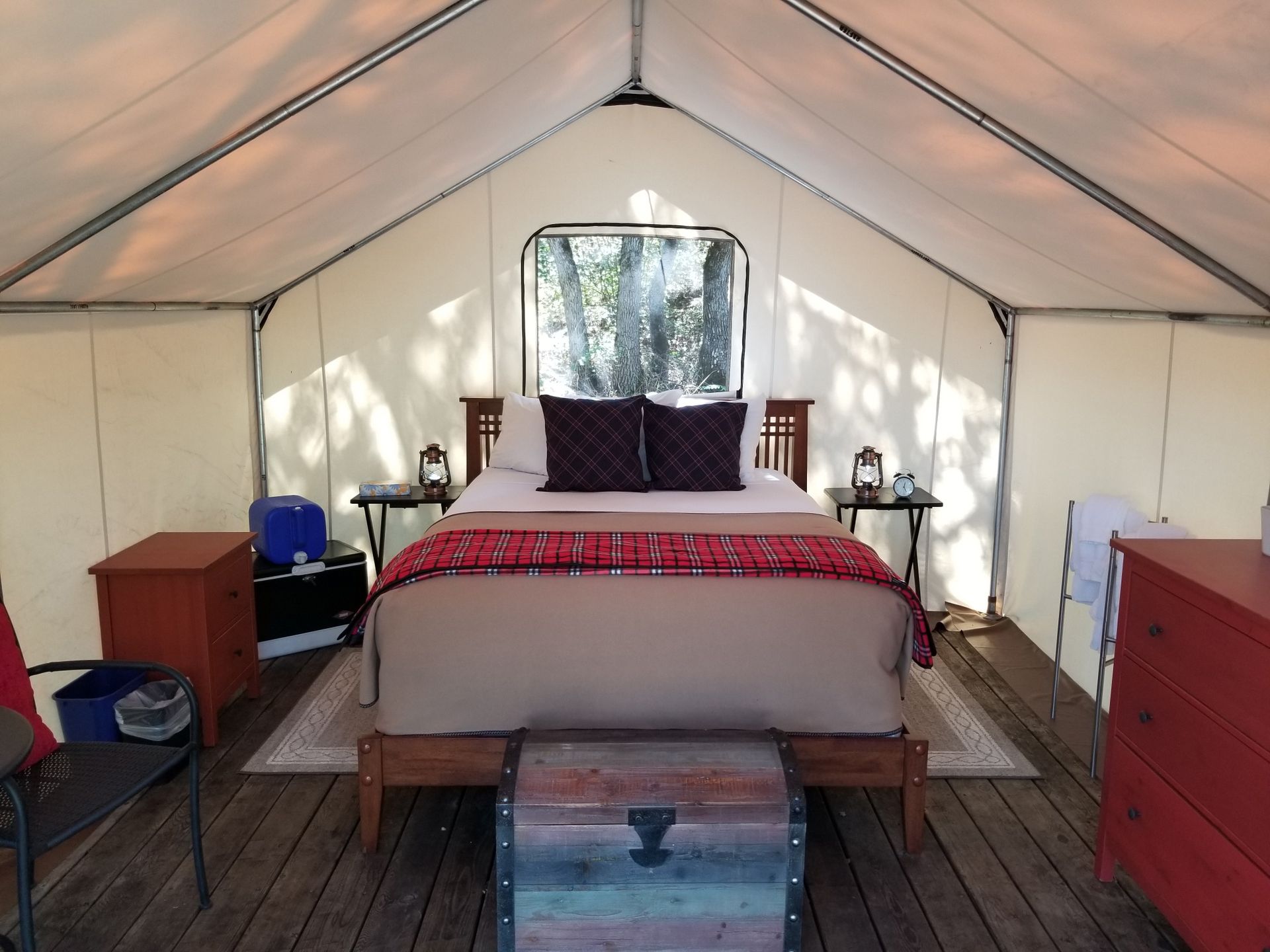 Inside a canvas tent with a bed, nightstands, and a trunk. Wooden floor and red furniture.