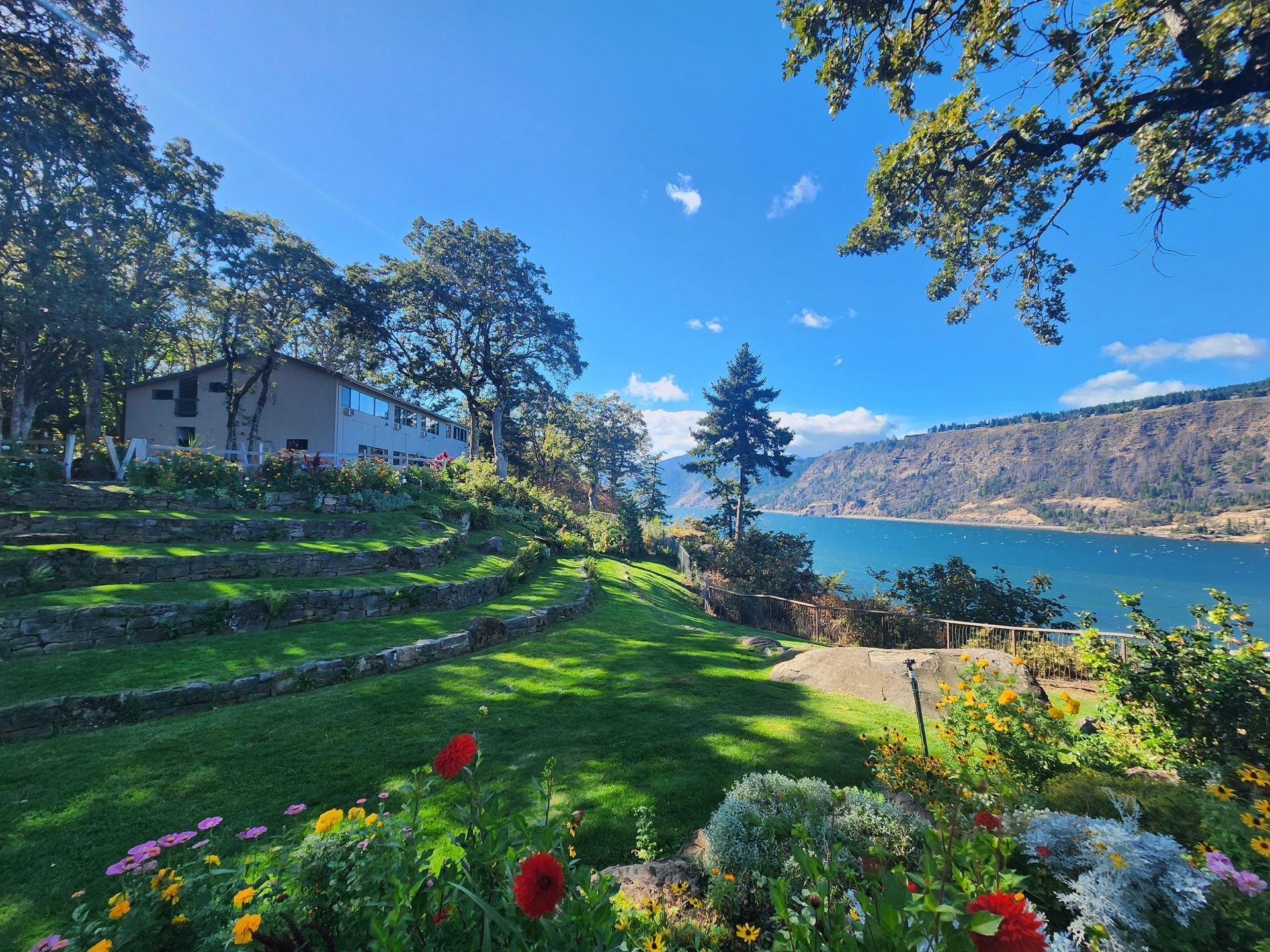 A terraced garden overlooking a lake, with a building on the left and mountains in the distance. Blue sky.