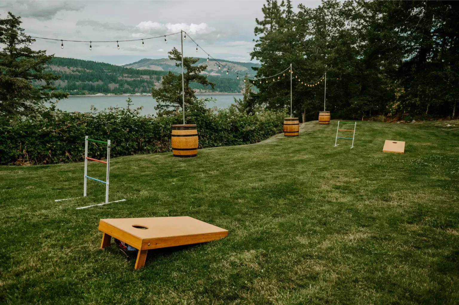 Outdoor games set up on a grassy hill overlooking a lake. Includes cornhole boards, ladder toss, and string lights.