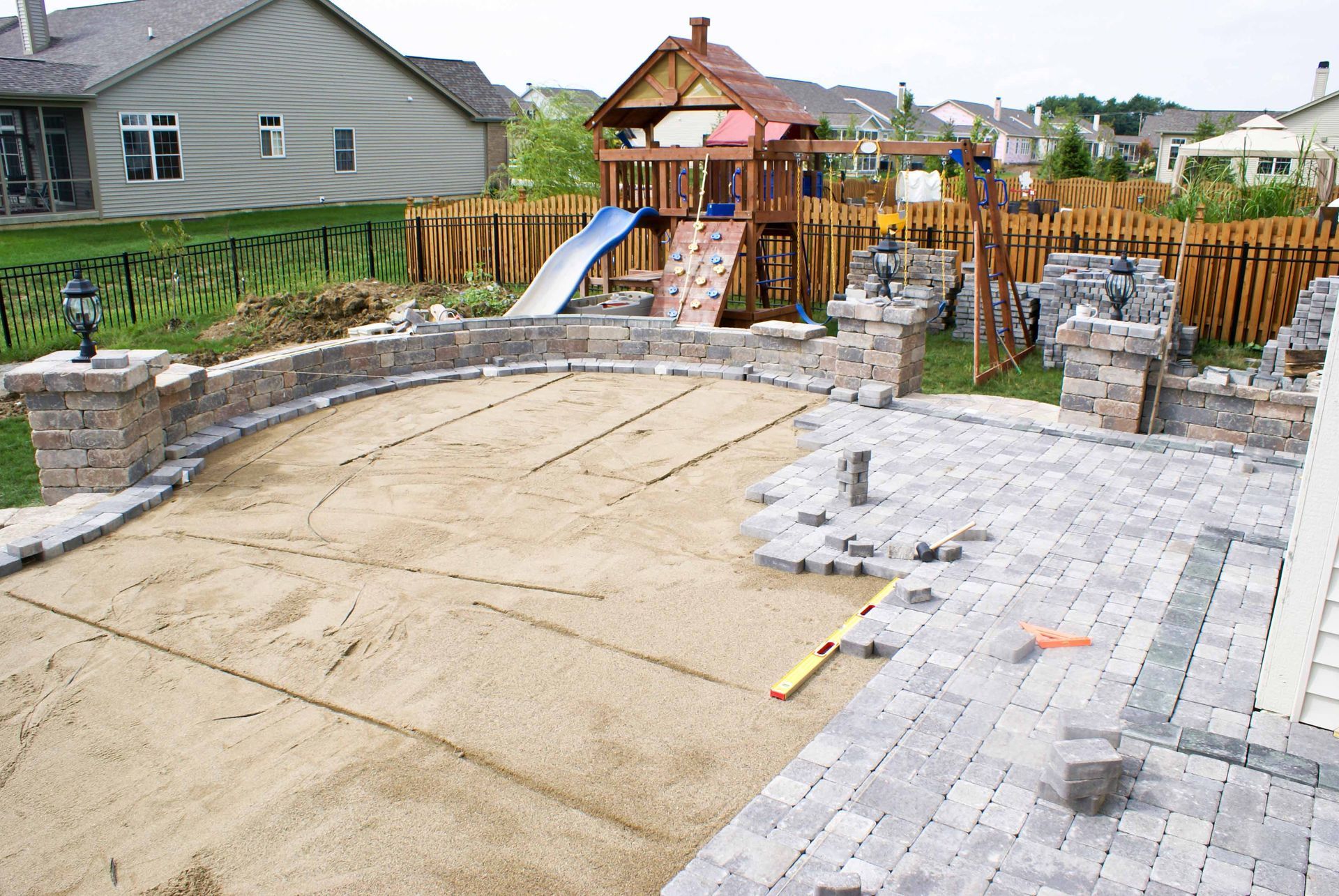 Backyard play area with curved sandbox, stone patio, and wooden playground set with slide and fence