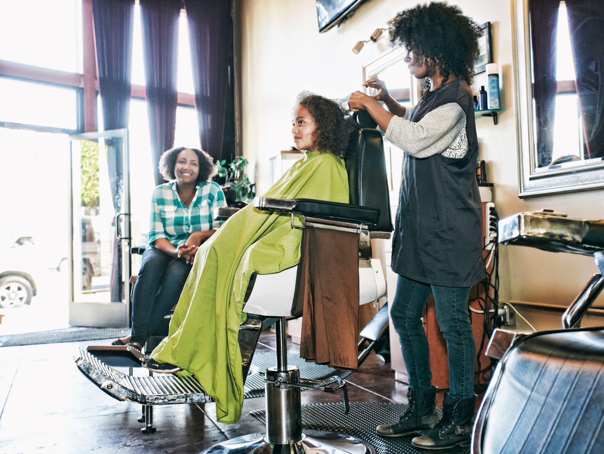 Woman styling a girl's hair in a salon. Another woman watches from a chair near an open door.